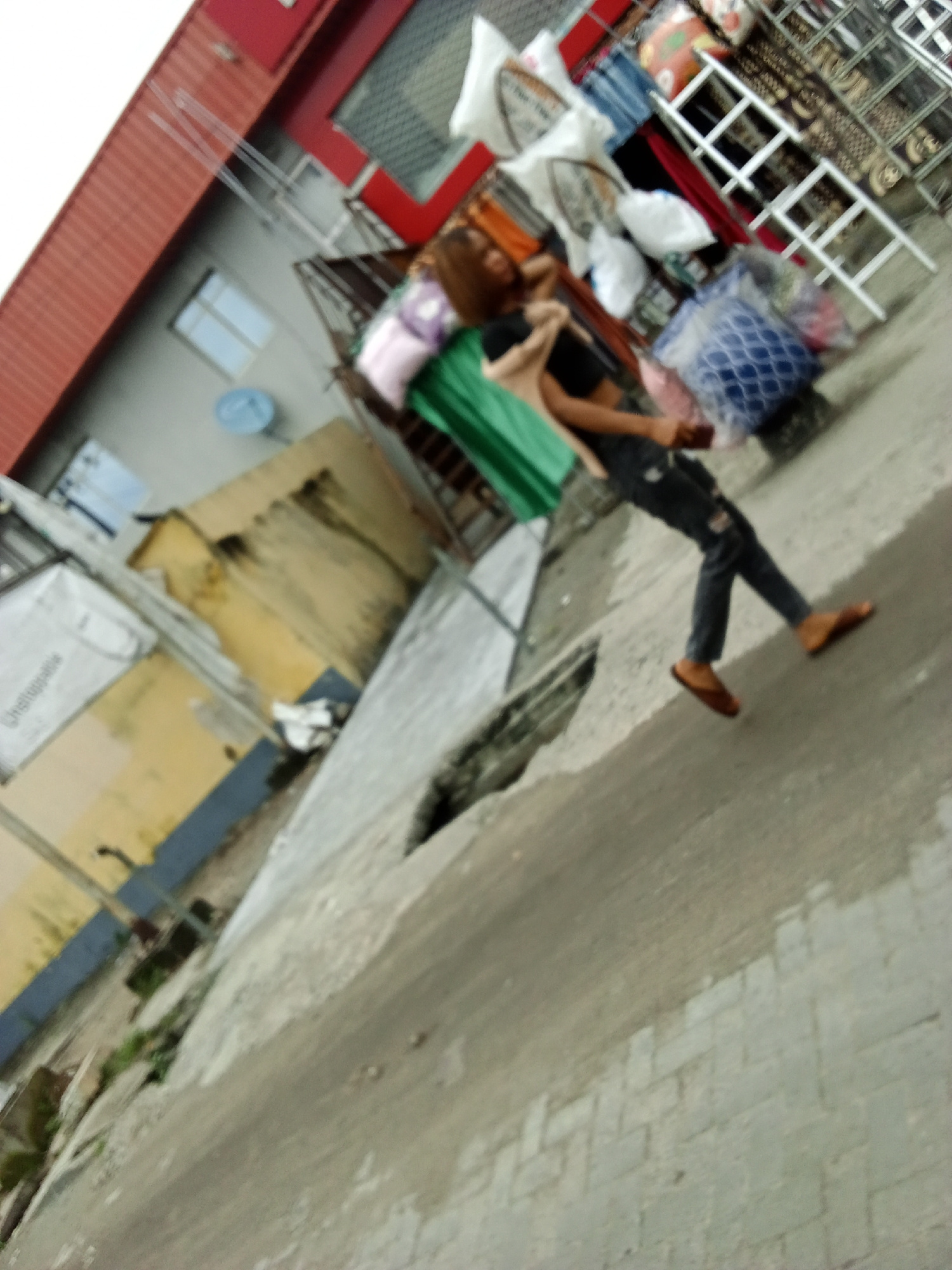 The media file depicts an outdoor scene with a person carrying several items. The person, whose face is blurred, is wearing dark pants and appears to be walking away from the camera. They are carrying a light-colored bag on their left shoulder and a green cloth draped over their right arm.
In the background, there are buildings and various objects. To the left, a yellow building with a blue section is visible, along with a satellite dish. To the right, there are what appear to be items for sale or storage, including bags and clothing stacked on shelves or racks. These items are diverse in color and texture. Some of the items are contained in blue and white patterned bags, and there are also some rolls of patterned material. The overall setting seems to be a street or an open area adjacent to buildings, possibly a market or a residential area with shops.
The ground is paved with concrete and some areas appear to be in disrepair, with cracks and uneven surfaces. The lighting suggests it is daytime, but the sky is not clearly visible. The weather appears to be clear. The image is blurry, making it difficult to discern specific details or emotions. There is no visible text in the media file.