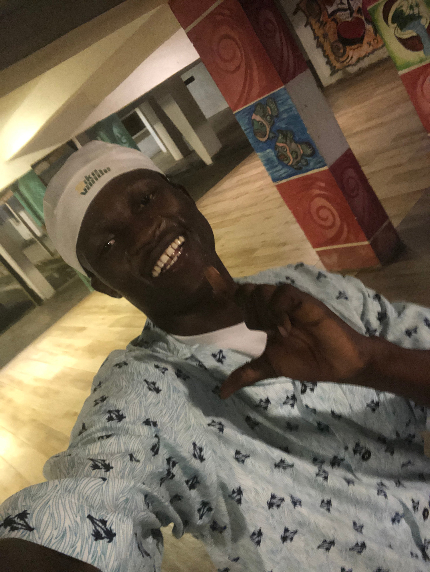 A man wearing a white cap and a light blue patterned shirt smiles at the camera, holding his hand up in a heart shape. He is in an indoor location with wooden floors and decorated pillars. The pillars have colorful patterns, including red swirls and blue sections with fish imagery. The lighting suggests it might be evening or an indoor artificial light setting. The overall impression is a casual, friendly portrait. There is no information in the image that places the scene specifically in Iwesolu, Nigeria.