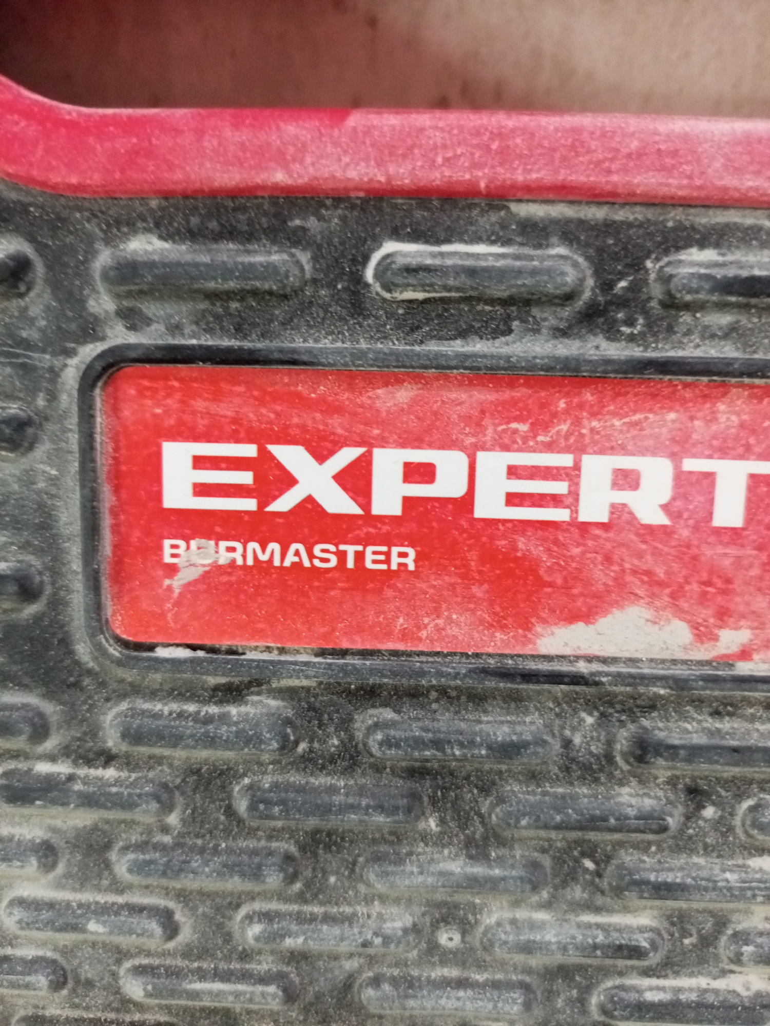 The image is a close-up, eye-level shot of a weathered, red and black decal on a textured surface, likely part of a tool or piece of equipment. The decal has white lettering, with the word "EXPERT" prominently displayed in larger font, followed by "BURMASTER" in smaller font below it. The red background of the decal is smudged and scratched, indicating wear and tear. The black textured background surrounding the decal features a pattern of raised, rounded rectangular shapes, also covered in a fine layer of dust or debris. The overall impression is one of a functional, outdoor, or workshop environment where the item has seen considerable use. There are no people or animals visible. The lighting appears to be natural and diffused, suggesting it could be daytime. The location context is Lymanka, Ukraine, but there are no specific visual cues in the image to confirm this.