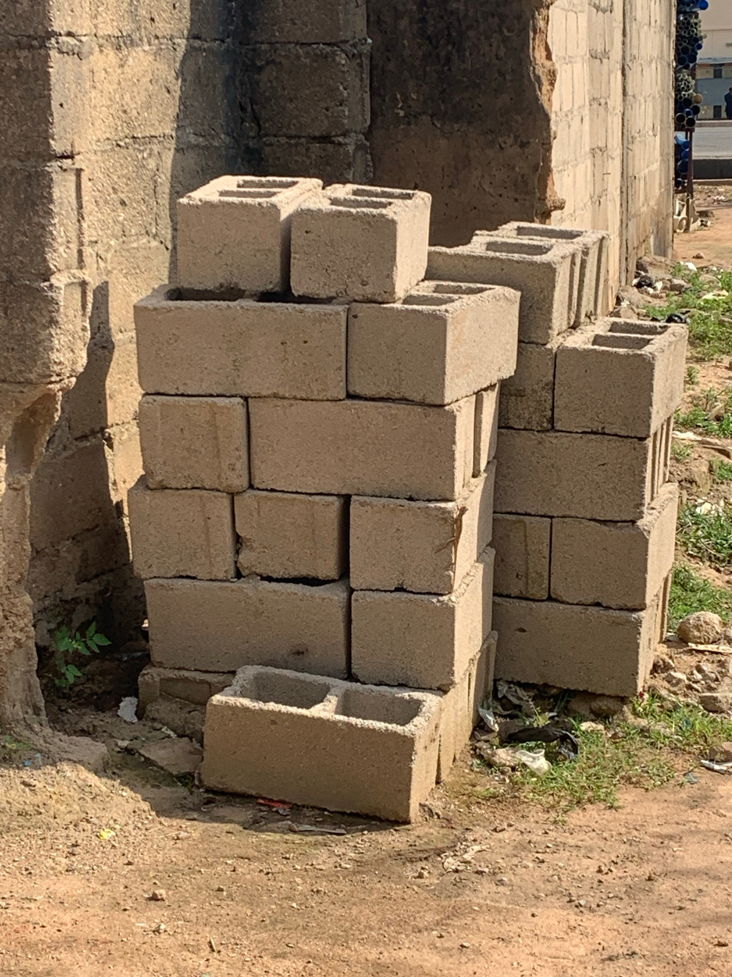 This image shows a pile of concrete blocks stacked on the ground. The blocks are of various sizes and appear to be standard construction blocks. Some of the blocks have holes running through them. The pile is situated on a dirt and gravel surface, with some patches of grass and weeds growing around it. In the background, there is a hint of a building with some pipes or containers stacked in front of it. The lighting suggests it is daytime, with shadows cast on the ground, indicating a sunny environment. The overall scene appears to be an outdoor area, possibly a construction site or storage area for building materials. There is no visible text in the image. No people are present. The scene is static, with no activity taking place.