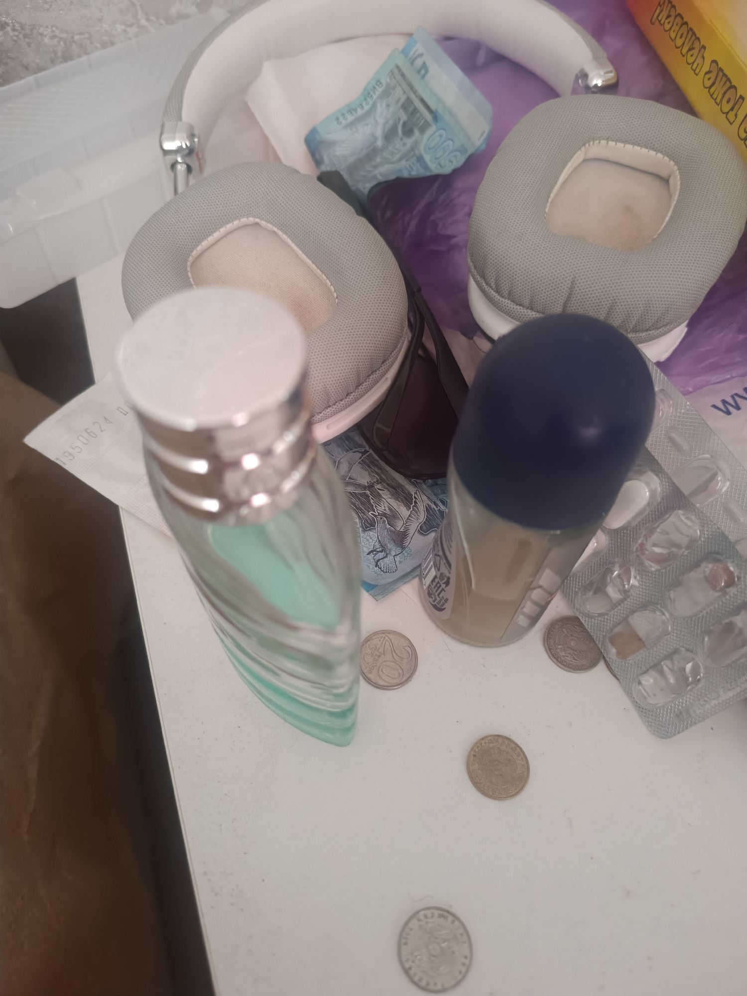 The image depicts a cluttered white surface with various objects. In the foreground, a tall, slender glass bottle with a light blue liquid and a silver cap is present. Next to it, a dark blue capped roll-on deodorant is visible. Scattered around these items are several coins, including one clearly showing "20". Behind these, a pair of light grey over-ear headphones with white accents lies on the surface. A partial blister pack containing white pills is also visible. Further back, a blue banknote with the numeral "500" is partially visible, along with a yellow box with text in a foreign language. The overall environment appears to be an indoor surface, possibly a desk or table, with hints of a textured grey background and a brown object in the lower left corner. There are no people or actions captured in the image.