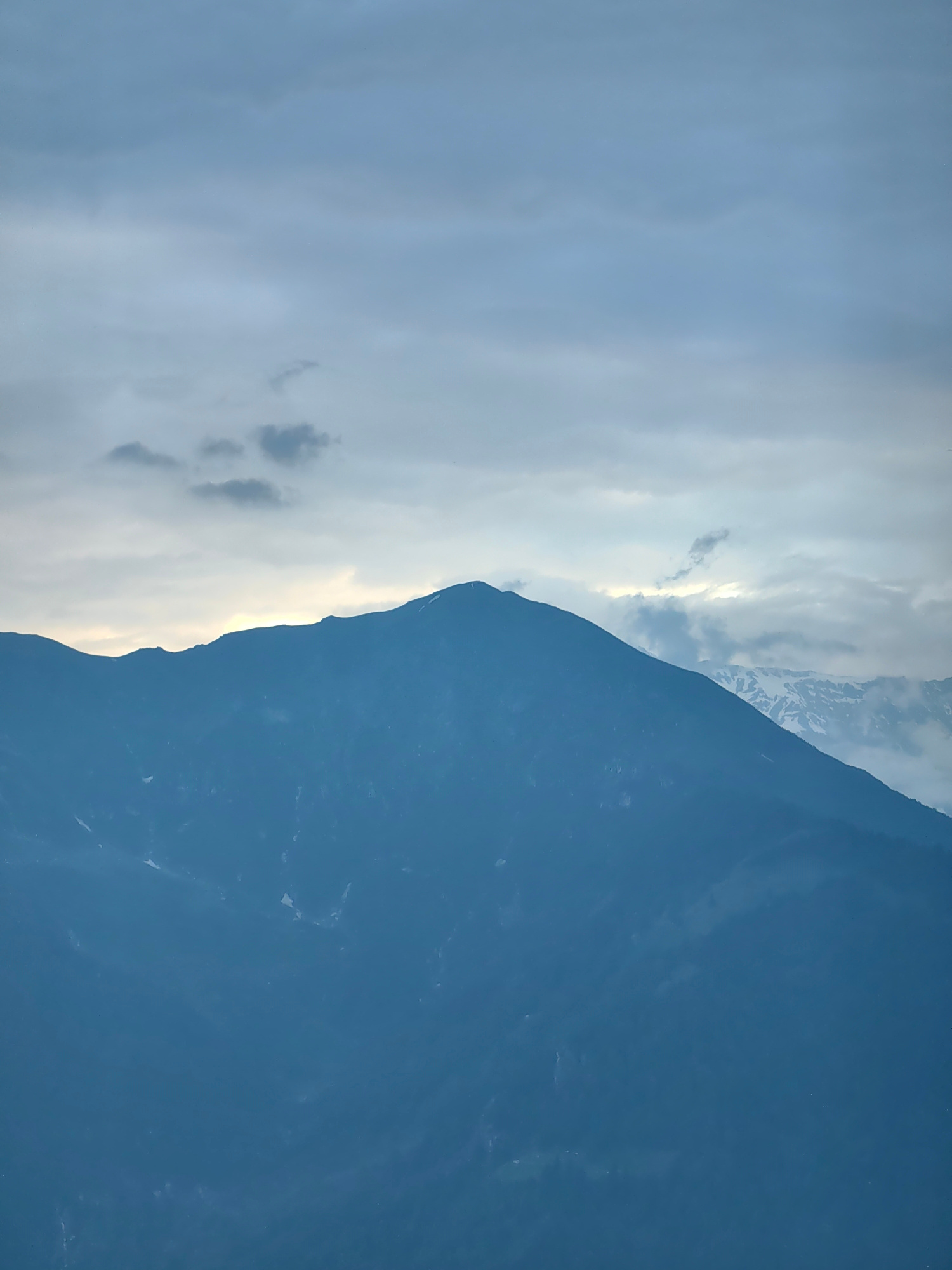 The image depicts a mountain range under a cloudy sky. The foreground features the silhouette of a large, dark blue mountain, its peak subtly outlined against the lighter sky. Some patches of snow are visible on its slopes. In the background, another, more distant mountain range is partially obscured by clouds, with some snow-capped peaks visible. The sky is filled with layers of gray and lighter blue clouds, with a bright, hazy band of light peeking through the clouds near the horizon, suggesting either sunrise or sunset. The overall mood is subdued and atmospheric, with the dark mountains creating a sense of scale and grandeur against the cloudy sky. The location is identified as Chhyal, India, implying a mountainous region within that area.