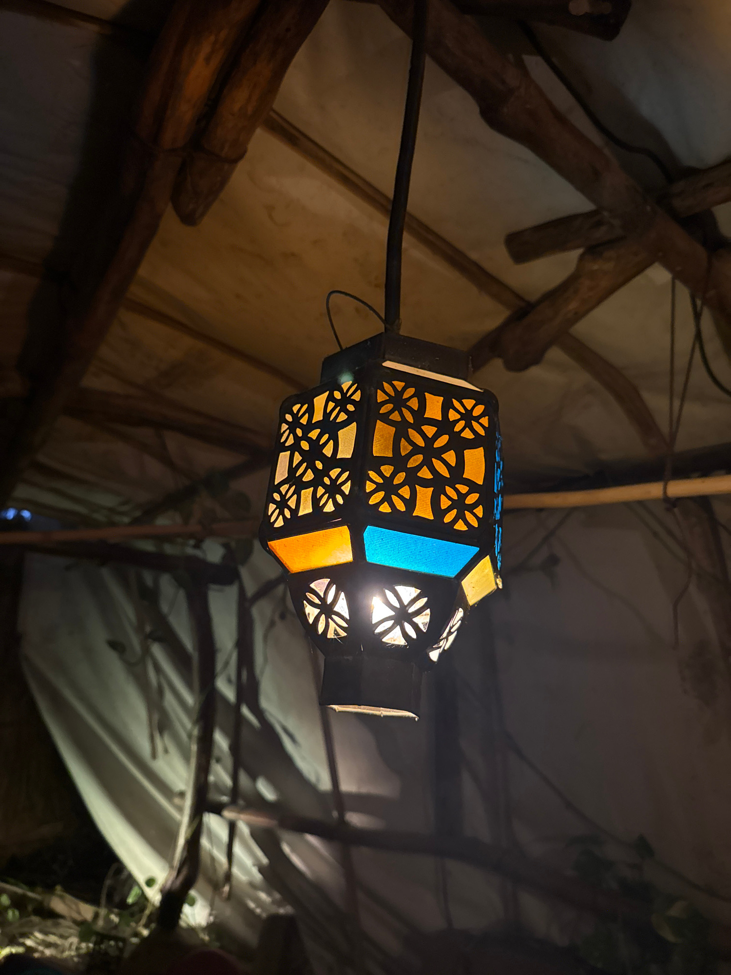 The image shows a decorative lantern hanging from a rustic wooden ceiling in what appears to be an outdoor or semi-outdoor setting. The lantern is hexagonal and features colored glass panels in orange and blue, interspersed with cut-out patterns. Light emanates from within the lantern, casting shadows and highlighting the intricate design. The ceiling is constructed from rough-hewn wooden beams and poles, supporting a translucent material that diffuses the ambient light. The background is dimly lit, with hints of fabric or tarpaulin and vegetation suggesting a somewhat makeshift or natural environment. The scene's atmosphere is warm and inviting, due to the lantern's glow. There are no people or animals visible in the image. The location is identified as Abang, Indonesia.