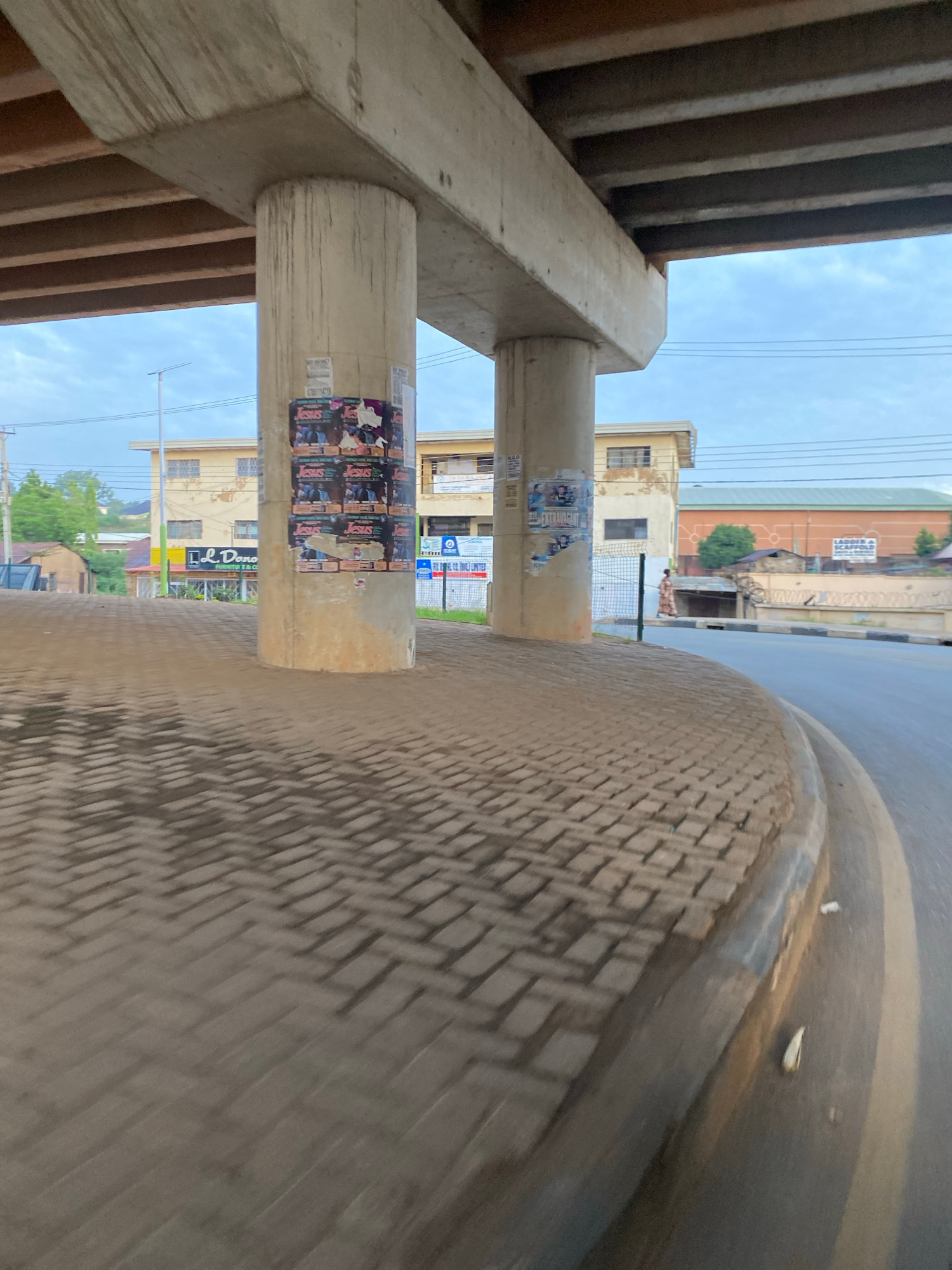 The image captures a view from under an elevated concrete structure, likely an overpass, in Jos, Nigeria. Two large concrete pillars support the structure, and the ground beneath is paved with interlocking bricks arranged in a herringbone pattern. Posters are affixed to the pillars.
In the background, several buildings are visible. To the left, a two-story building with a cream-colored facade and dark-framed windows stands. Adjacent to it, a building with a yellow facade features a sign that reads "L.L. Dono FURNITURE & CO." Further to the right, another building with a similar cream-colored facade is present. Beyond these, a larger building with a distinctive orange exterior and dark architectural details is situated. A sign on this building reads "LADDER A SCAFFOLD."
The sky is overcast with a pale blue hue. Power lines are strung across the upper portion of the scene. A paved road curves to the right, with a curb separating it from the brick-paved area. A person in a long, patterned dress is walking on the side of the road. The scene suggests a somewhat developed urban environment.