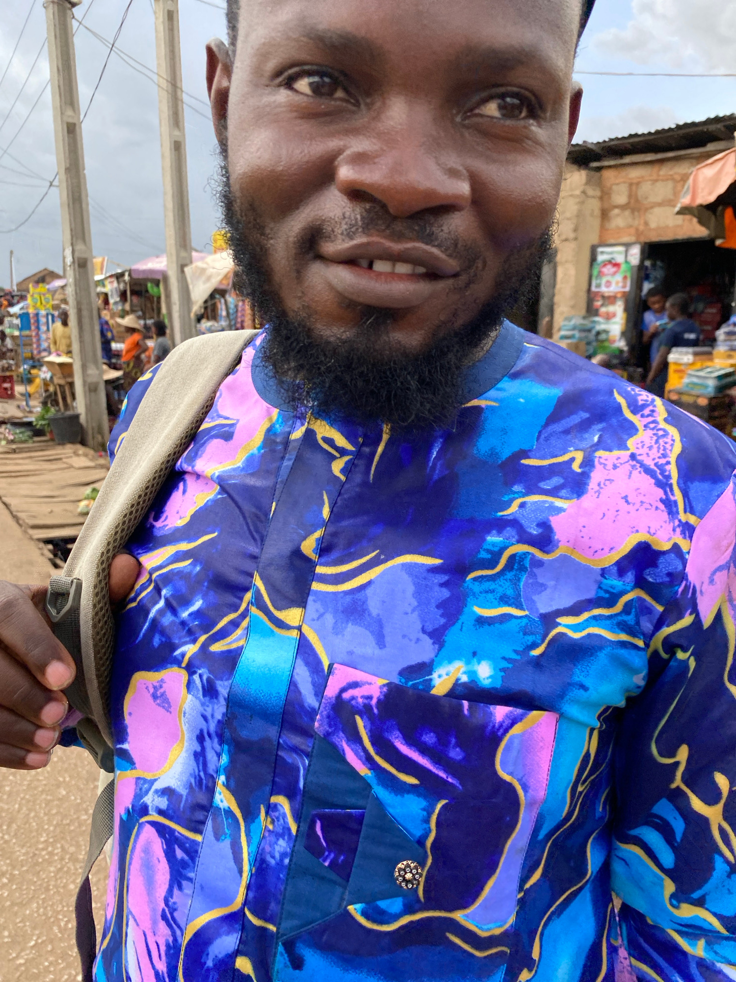 This is a close-up, vertically oriented, medium shot of a Black man smiling with his head tilted. He is wearing a vibrant, blue, purple, and pink patterned shirt with yellow accents. The shirt has a dark blue collar and a small, ornate, gold button on the right side of his chest. A portion of a beige backpack strap is visible crossing his chest.
The man's face is turned towards the camera, and he has a short, dark beard. His left eye is clearly visible, and he appears to be in good spirits, with a wide smile revealing his teeth.
In the background, out of focus, are elements of an outdoor marketplace. We can see stalls with various goods, some of which appear to be food items. There are also indications of people shopping and milling about, suggesting a busy day. Overhead, there are power lines and poles, common in urban or semi-urban environments. The sky appears overcast, suggesting it might be a cloudy day. The overall impression is a candid, everyday moment captured in a lively setting.