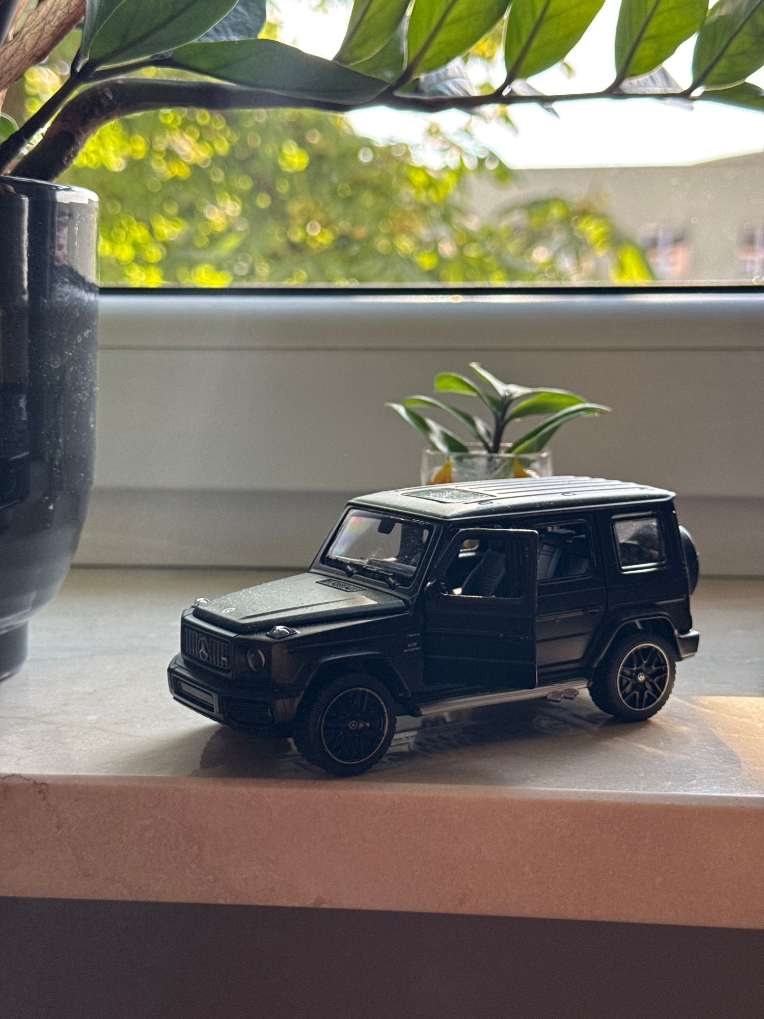 A black toy Mercedes-Benz G-Class is positioned on a windowsill. The toy car has its driver's side door open. In the background, a plant in a glass vase is visible, with out-of-focus greenery and buildings seen through a window. A portion of a dark pot is on the left side of the frame. The windowsill appears to be made of a speckled, light-colored material. The scene is lit by natural light, casting a warm glow on the car and windowsill. The location is not specified and cannot be determined from the image.
