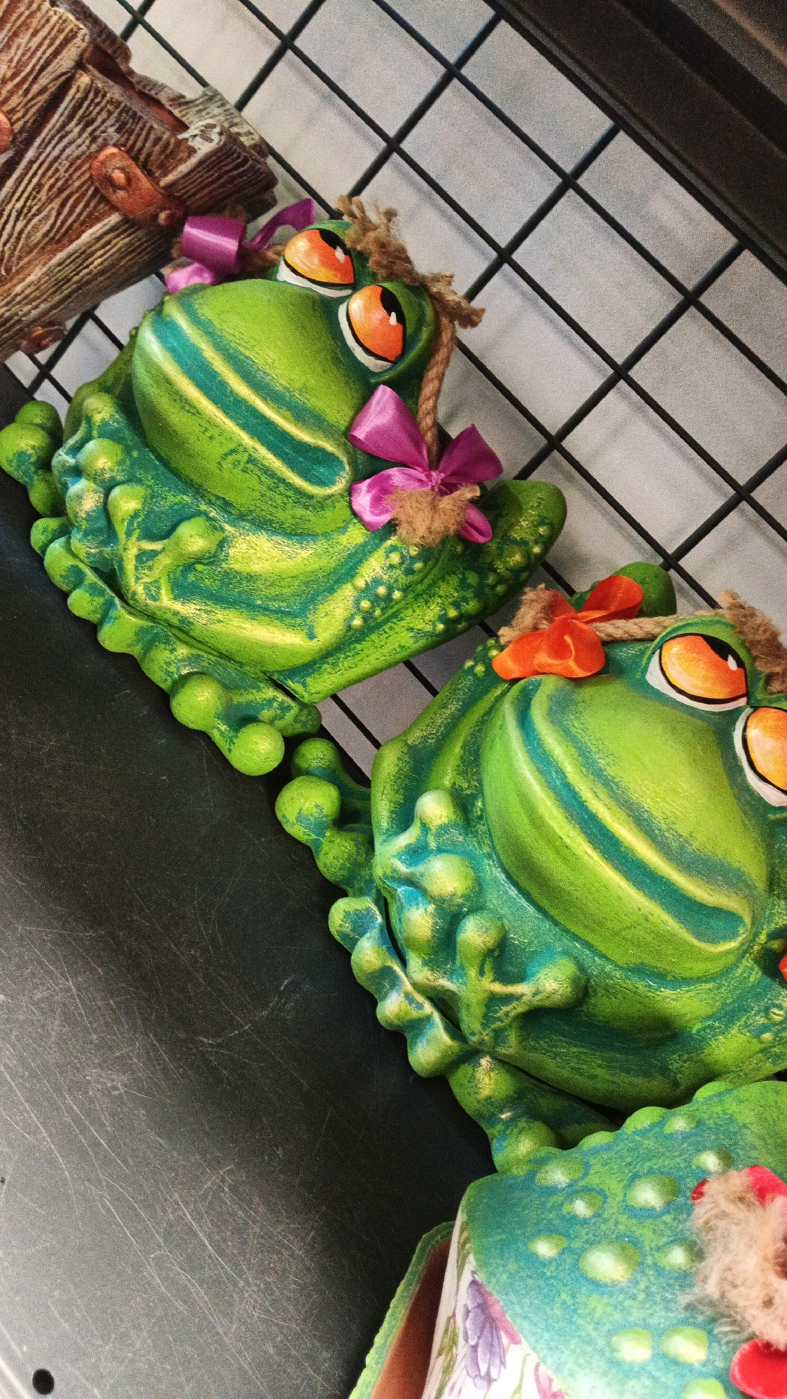 Two decorative ceramic frogs are displayed on a dark, textured surface, possibly a shelf or tabletop. The frogs are bright green with yellow and blue accents, and they have large orange eyes. They are adorned with small bows and what appears to be twine or rope around their heads, giving them a whimsical appearance. The background consists of a black wire grid. The image is taken from a slightly elevated angle, looking down at the frogs. There is no indication of the city of Kryvyi Rih, Ukraine, in the visual information provided.