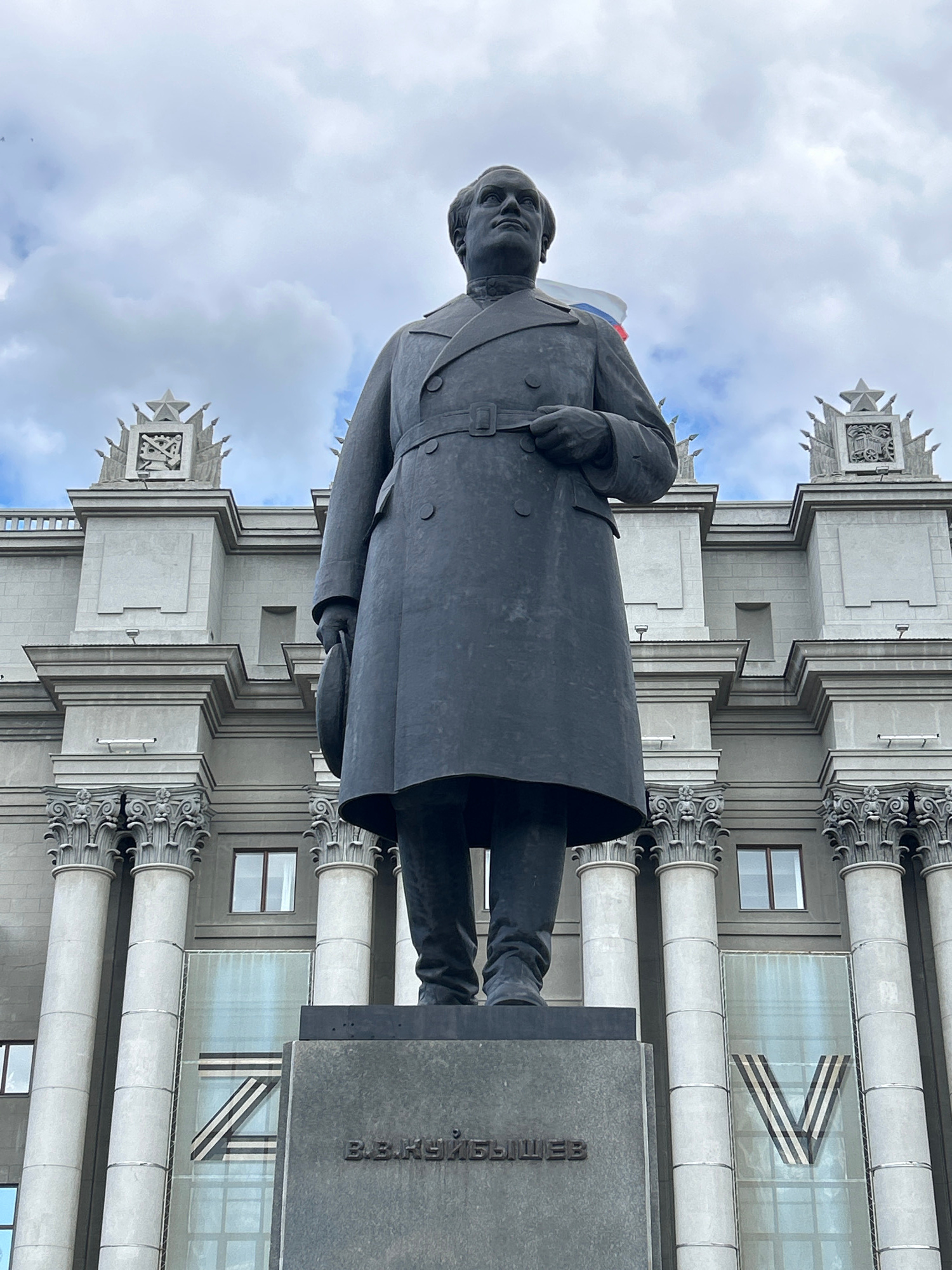 This media file features a large, dark-colored statue of a man in a coat. The statue is positioned prominently in the foreground, facing slightly to the left. Behind the statue is an imposing, light-colored building with classical architectural elements, including columns with ornate capitals and decorative pediments. The building has large windows, one of which displays a graphic of a large, stylized "V" symbol made of dark stripes against a lighter background. The sky above is cloudy and overcast, suggesting a cool or temperate day.
The statue appears to be made of bronze or a similar dark metal and has a solemn expression. A small Russian flag is visible draped over the shoulder of the statue. The base of the statue is a dark, granite-like material with visible text engraved on its side. The text, in Cyrillic script, appears to read "ВЕЛИКИЕ". The overall impression is one of gravitas and historical significance, likely commemorating a notable figure. The building's architecture and the presence of the statue suggest a public square or a place of historical importance in Samara, Russia. The time of day is difficult to ascertain due to the overcast conditions, but the lighting suggests it is daytime. The emotions evoked are a sense of respect and perhaps awe.