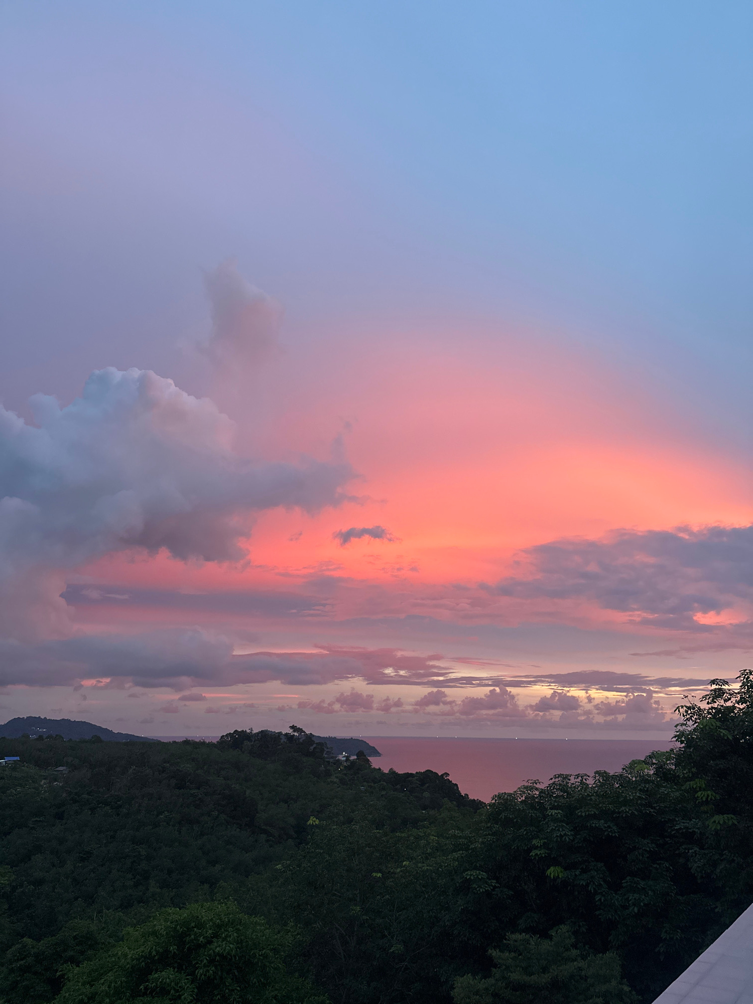 This image captures a dramatic sunset over the ocean, viewed from a hillside in Kamala, Thailand. The sky is a vibrant spectacle of color, transitioning from a soft blue at the top to a vivid pink and orange, illuminated by the setting sun. Wispy clouds, tinged with shades of grey, pink, and orange, are scattered across the sky.
Below the sky, a lush, dark green forest covers the undulating hills. The trees are dense and appear to be tropical foliage, creating a stark contrast with the bright sky. In the distance, the calm ocean stretches towards the horizon, reflecting the colors of the sunset. The water appears calm, with a few faint lights visible on the surface, possibly from boats or distant shorelines. The overall impression is one of serene natural beauty at dusk.