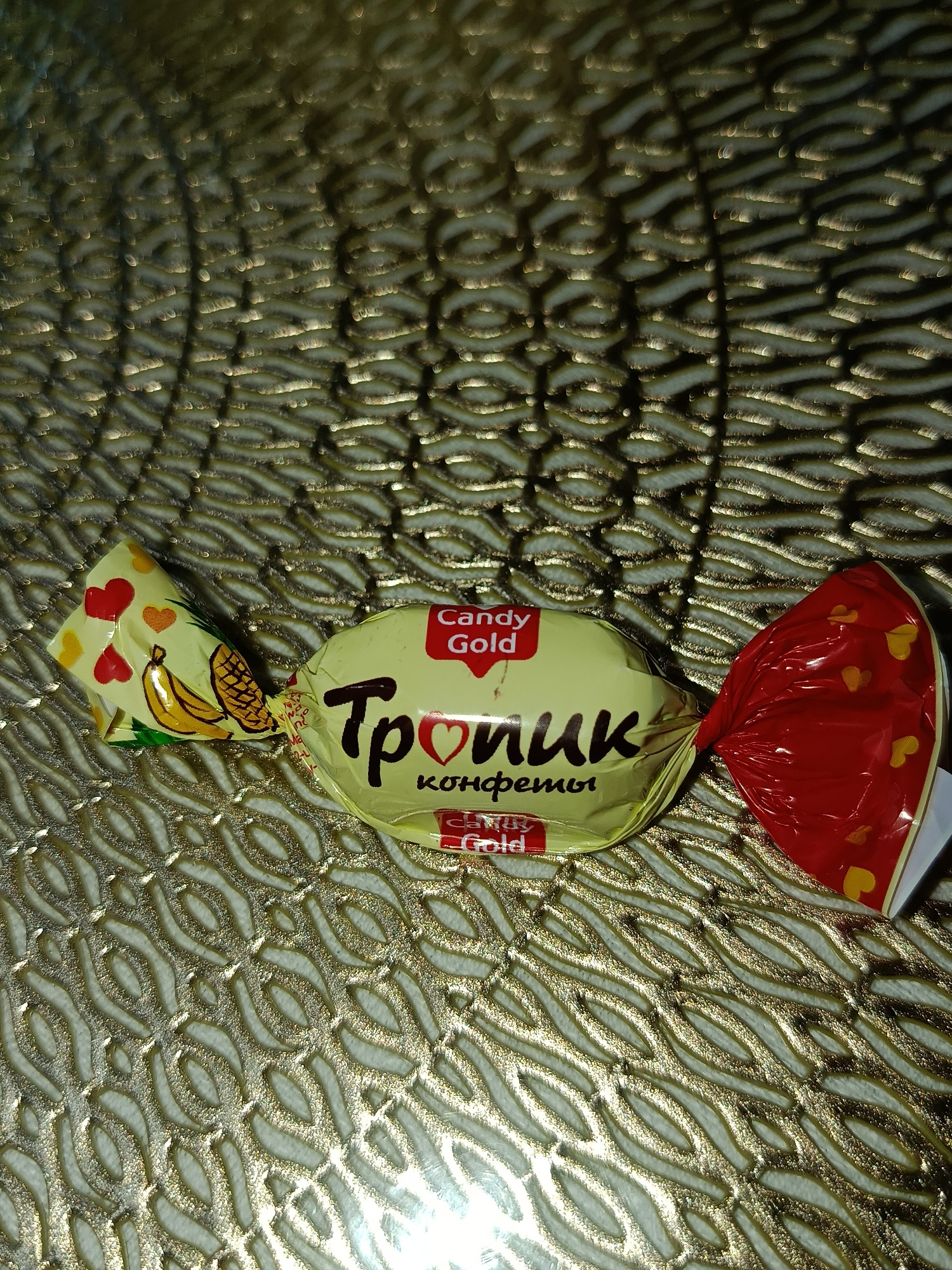 The image shows a single candy wrapper lying on a textured, golden surface. The wrapper is primarily yellow with red accents and features illustrations of bananas, pineapples, and hearts. The text on the wrapper is in Cyrillic and English, with visible words including "Candy Gold" and "Тропик". There are no people or identifiable landmarks present in the image, and no information suggests it is located in Tashkent, Uzbekistan.