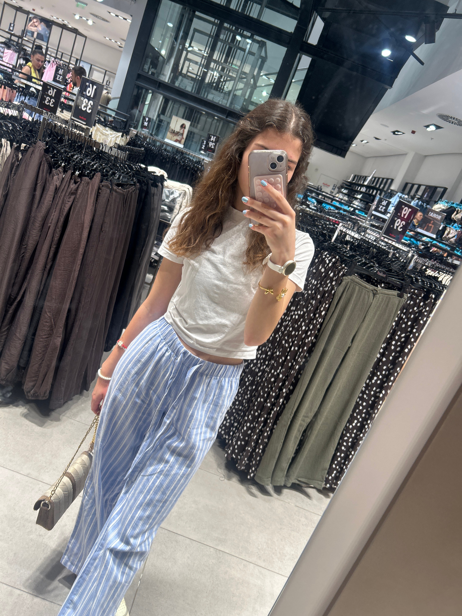 A young woman is taking a mirror selfie in a clothing store. She is wearing a white t-shirt and light blue and white striped pants. She is also holding a beige and white patterned purse. In the background, there are racks of clothes and a large mirror reflecting the store. The store appears to be in Ruse, Bulgaria, based on the "RUSE" text visible on the price tags in the background.