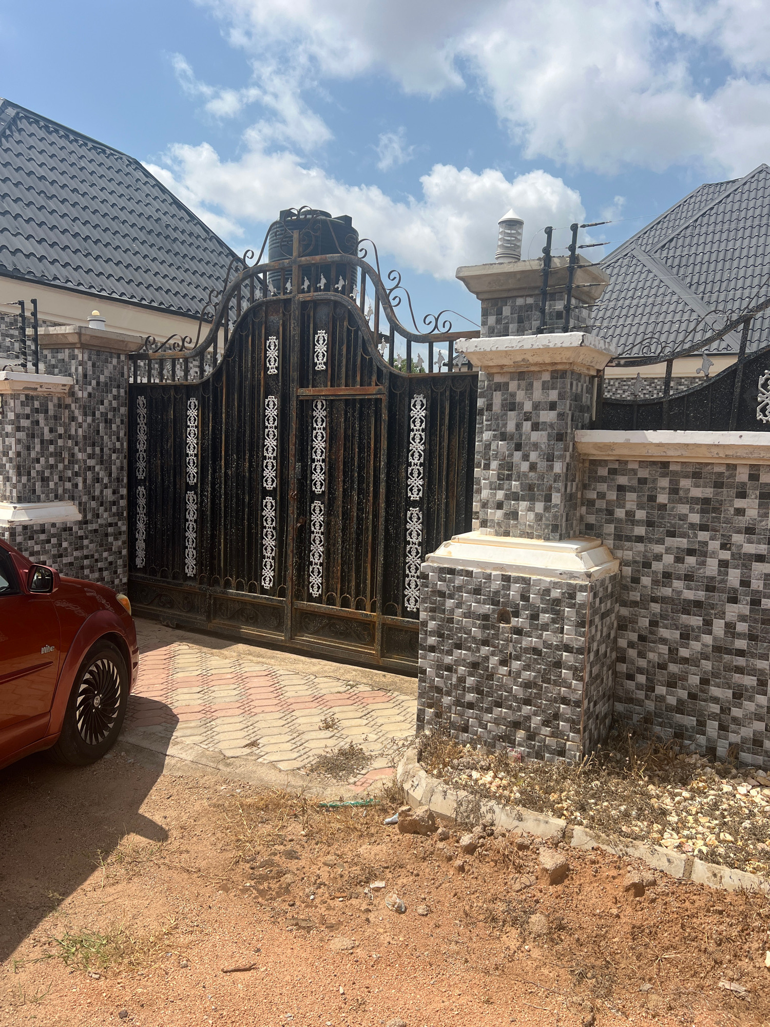 The image depicts an outdoor scene in Howayi, Nigeria, under a bright, partly cloudy sky, suggesting it is daytime. The primary subjects are a dark metal gate, part of a building's facade, and a portion of a red car.
The gate is ornate and appears to be made of wrought iron with decorative elements. It is set against a wall covered in small, square mosaic tiles in shades of grey and white. Behind the gate, a tiled walkway is visible. The building features a dark, wavy-patterned roof.
To the left of the gate, a concrete pillar is visible, topped with a cylindrical object and what appear to be electrical insulators or components. The ground surrounding the tiled area is sandy and dry, with sparse vegetation. The red car is parked near the gate, with its front wheel and a section of its body visible. The car has a distinctive wheel design.
There are no people visible in the image. The overall impression is of a residential or commercial property entrance in a warm, possibly arid climate, given the sandy ground and dry vegetation. The scene is static, with no specific activity taking place. No visible text is present in the image.