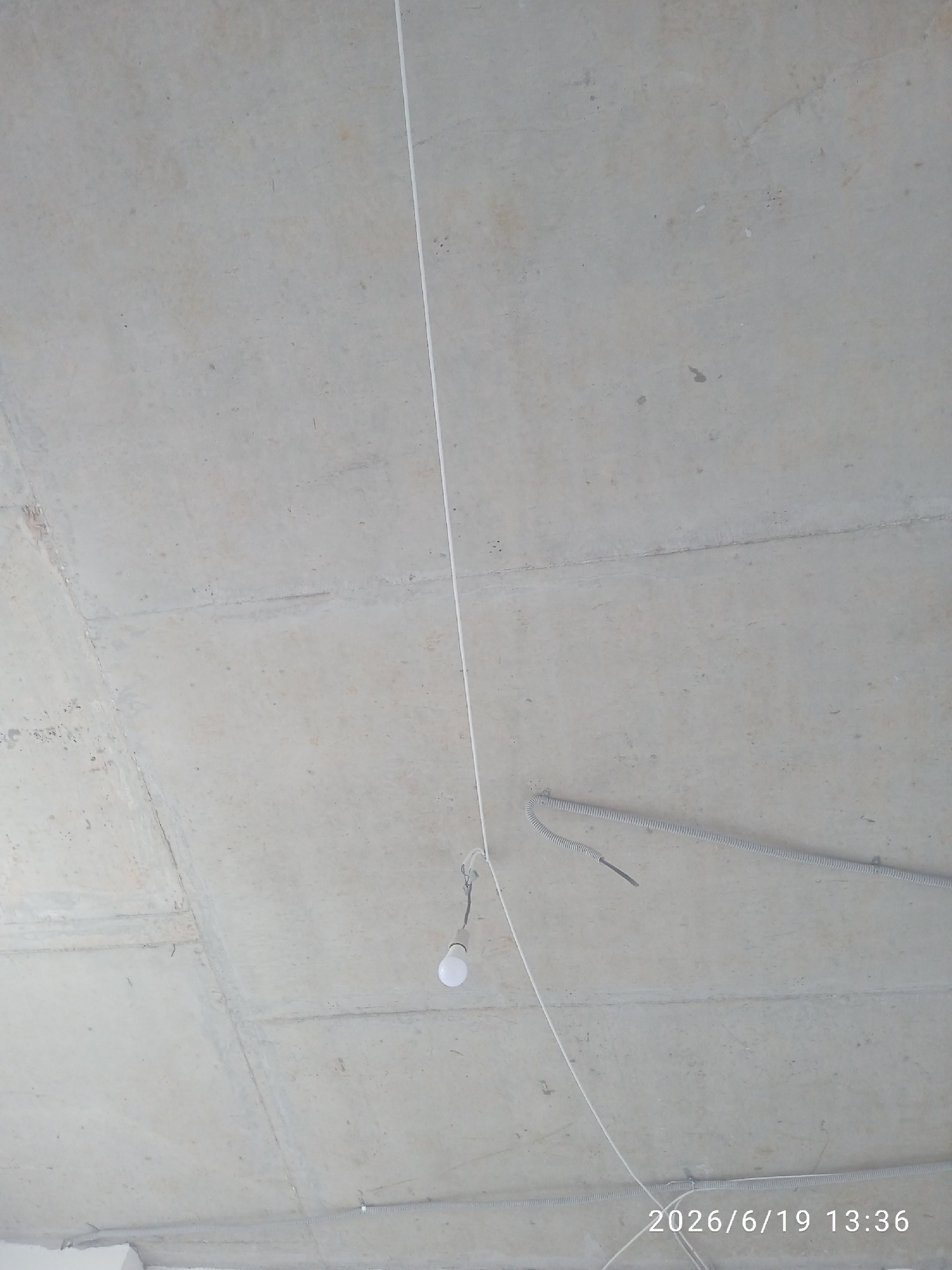 The image shows a concrete ceiling with electrical wiring and a light bulb. There are two lengths of gray corrugated conduit attached to the ceiling, one running horizontally across the middle of the frame and another lower in the frame. A white electrical wire hangs from the ceiling, with a single light bulb attached to it. The date and time stamp "2026/6/19 13:36" is visible in the lower right corner of the image. The scene appears to be an unfinished interior space, possibly a building under construction. There are no people visible in the image. This image was taken in Zvenigorod, Russia.