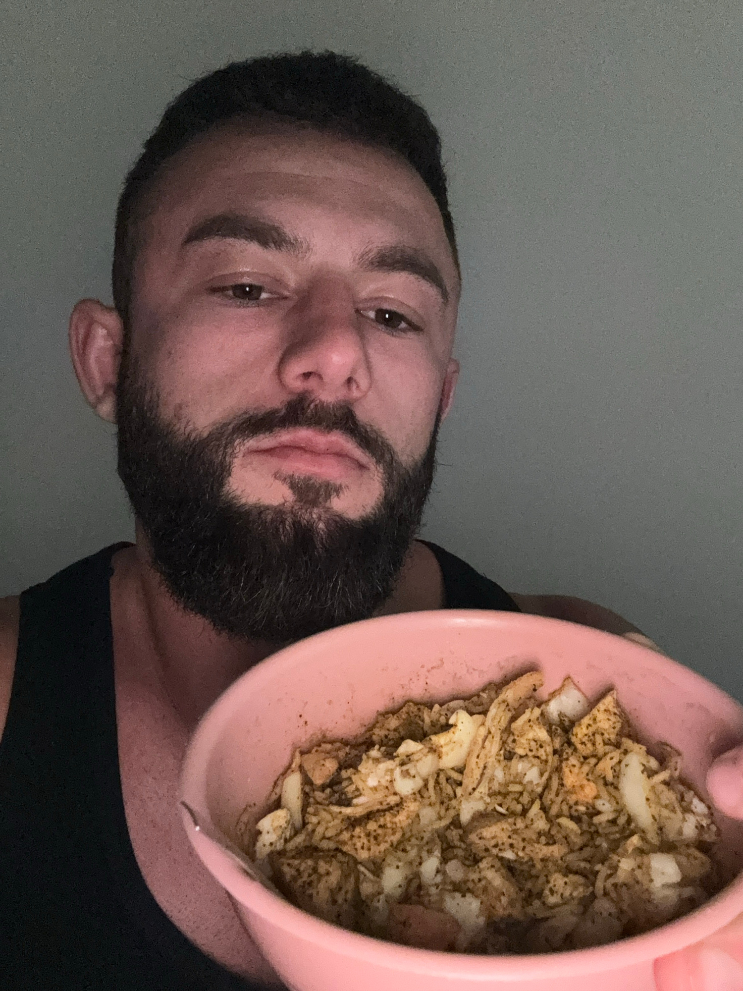 A close-up, vertically oriented photograph (originally rotated 90 degrees counter-clockwise for the viewer) captures a man holding a vibrant pink bowl of food. The man, positioned on the left side of the frame, has dark hair, a well-groomed dark beard, and a mustache. He looks directly at the camera with dark eyes and a neutral, slightly pensive expression, his lips subtly pursed. He is wearing a dark, sleeveless top.
The pink bowl, held by a hand partially visible at the top right, contains a meal primarily composed of shredded or diced meat, likely chicken, mixed with cooked onions and possibly grains. The food is heavily seasoned with dark spices, giving it a speckled appearance. The background is a plain, light grey wall, indicating an indoor setting. The lighting is even and moderate. No text is visible in the image. This scene takes place in Sofia, Bulgaria.