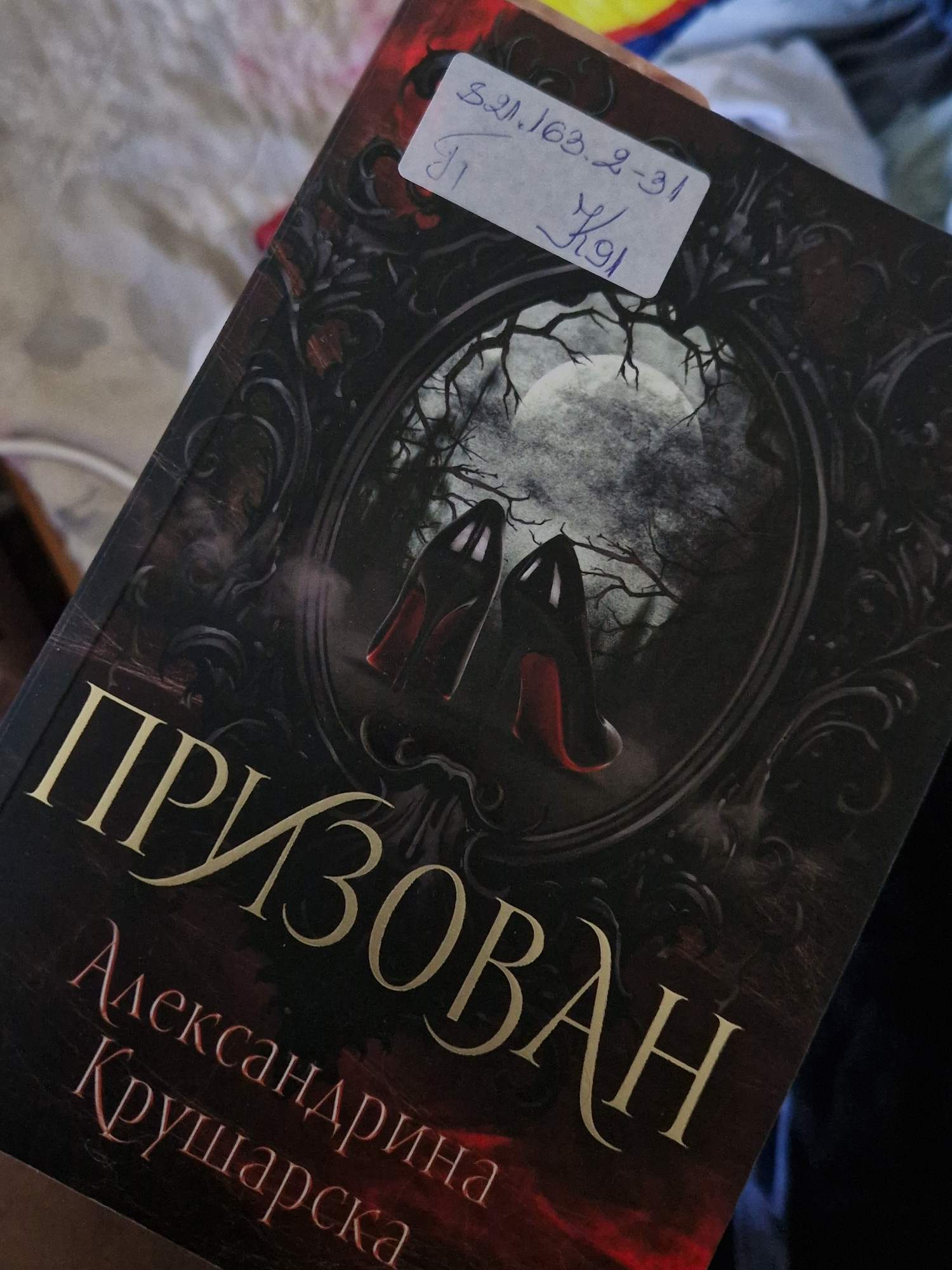 The image shows the cover of a book titled "Призван" (Prizvan) by Александрина Крушарска (Alexandrina Krusharska). The book cover features a dark, gothic design with a circular illustration in the center depicting a full moon, silhouetted trees, and two black high-heeled shoes with red soles. A white sticker with handwritten text, "821.163.2-31" and "K91", is affixed to the upper right portion of the cover. The book is held at an angle, obscuring part of its lower section. The background appears to be a soft, patterned fabric, possibly a blanket or sheet, in muted colors. There is no visible indication of the city of Raduvene, Bulgaria, in this image.
