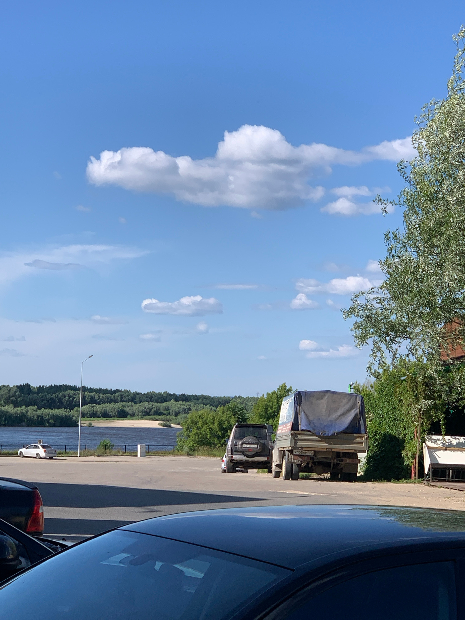 The image depicts a waterfront scene on a sunny day, likely in Pavlovo, Russia. The sky is a clear blue with scattered white clouds. In the foreground, the dark reflective surface of a car occupies the right side of the frame. Beyond the car, a paved area or road is visible, with a few parked vehicles. A prominent subject is a trailer truck covered with a blue tarp, parked on the side of the road. Beside it, a dark SUV is also parked.
The background reveals a lush green treeline running along the edge of a wide body of water, presumably a river or lake. The water appears calm. A solitary lamppost stands near the water's edge. The presence of trees, water, and a road suggests an outdoor, possibly recreational or industrial, area near a natural landscape. The bright sky and clear visibility indicate it is daytime, likely midday or afternoon, with fair weather. No specific activities or emotions are explicitly conveyed, but the scene suggests a peaceful, possibly transient moment by the water.
There is some visible text on the trailer truck, though it is partially obscured and difficult to read clearly. A license plate on the SUV reads "V155HH 152".