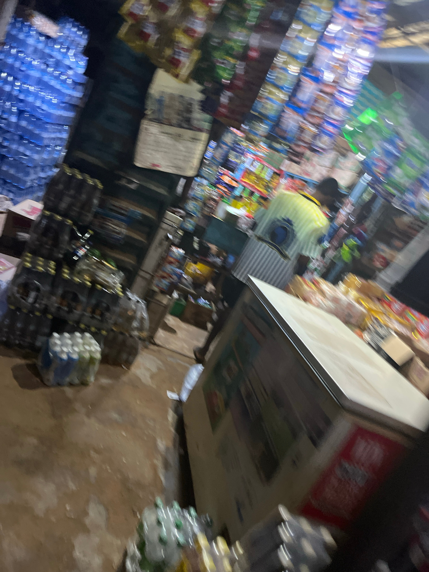 The image depicts a dimly lit convenience store interior in Unknown, Unknown. Stacks of bottled drinks and packaged goods are visible on shelves and the floor. A person wearing a yellow and white striped shirt is partially visible in the center, appearing to be working or shopping. The store seems to be cluttered with merchandise.