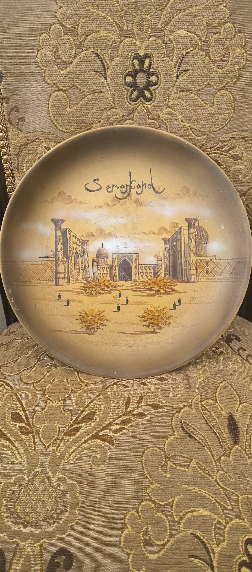 A circular, decorative plate sits on a patterned fabric background. The plate features a hand-painted scene depicting an architectural complex with multiple domed structures and tall, ornate pillars, characteristic of Central Asian or Middle Eastern design. The word "Samarkand" is written in a flowing script above the artwork. The fabric beneath the plate has a damask pattern in shades of gold and brown. The lighting suggests an indoor setting.