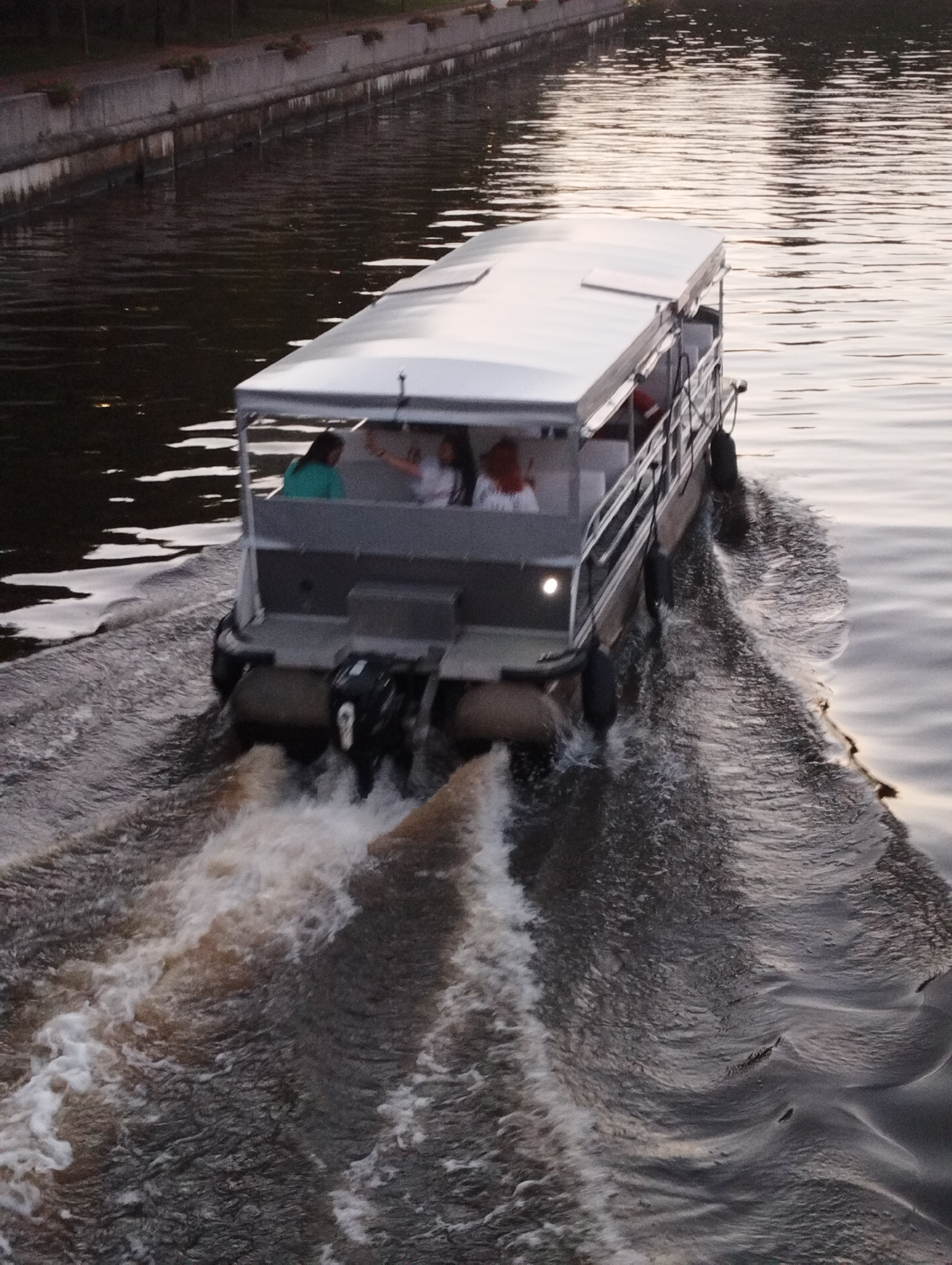 The image shows a pontoon boat moving through water, leaving a wake. The boat has a white canopy covering its top, and railings along its sides. Several people are visible on the boat. The boat is moving away from the viewer, towards the right of the frame. The water is dark and rippled, reflecting the sky. In the background, there is a concrete embankment with some vegetation on top, suggesting an urban setting, possibly a canal or river within Kaliningrad, Russia. The lighting indicates it is either early morning or late evening.