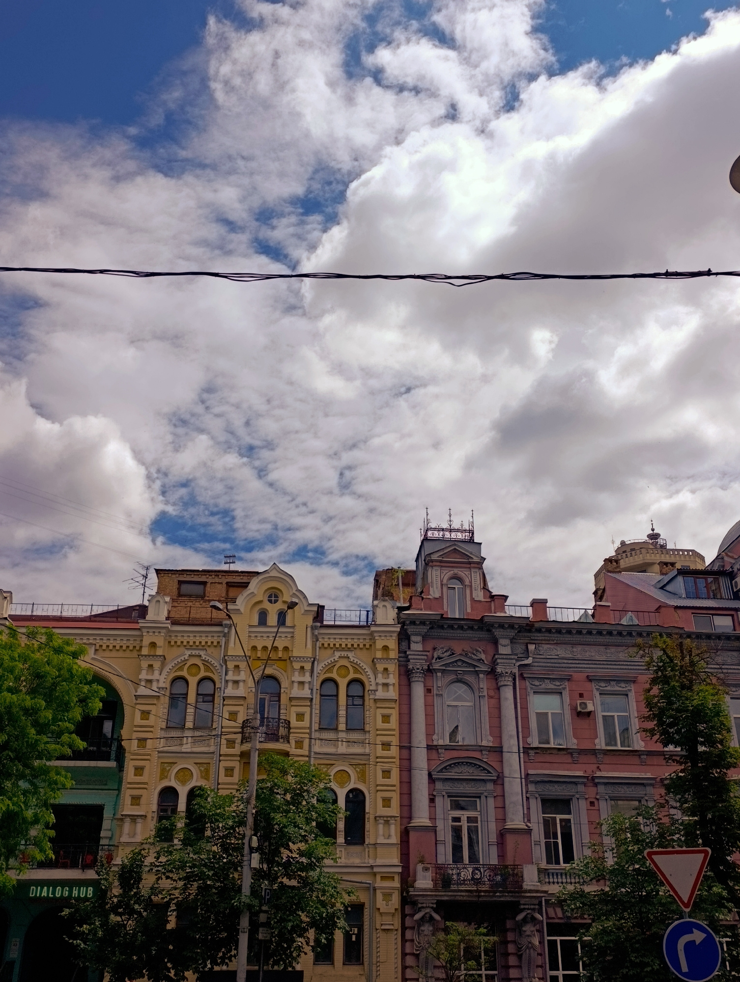 This is a ground-level, eye-level, slightly upward-looking, daytime, medium shot photograph taken in Kyiv, Ukraine. The image features a street view of historical buildings with ornate architectural details. The sky above is filled with dramatic, fluffy white clouds against a bright blue backdrop. A dark power line runs horizontally across the upper portion of the frame.
The buildings are multi-story structures with distinct facades. The building on the left is painted a light yellow with intricate white decorative elements, arched windows, and balconies. The building to its right is painted a pinkish-mauve color, also featuring detailed architectural embellishments, columns, and windows. Trees with lush green foliage partially obscure the lower portions of the buildings.
In the lower left corner, a green sign with white text that reads "DIALOG HUB" is visible, suggesting a commercial establishment. Street signs, including a white triangular sign with a red border and a blue circular sign with a white arrow indicating a right turn, are present in the lower right portion of the image. There are no people visible in the frame. The overall impression is one of an urban street scene in a European city, highlighting the juxtaposition of historical architecture and modern infrastructure.