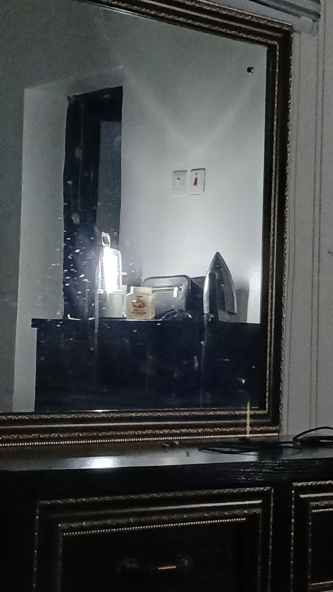The image shows a reflection in a mirror of a dresser with several items on top. The dresser is dark and ornate, with multiple drawers and decorative trim. On the dresser are a lamp, a bag or purse, and what appears to be a bottle with a label. To the right of these items, a black iron is standing upright. The reflection also shows a wall with two light switches and a doorway. The overall scene suggests an indoor setting, possibly a bedroom or dressing room.
