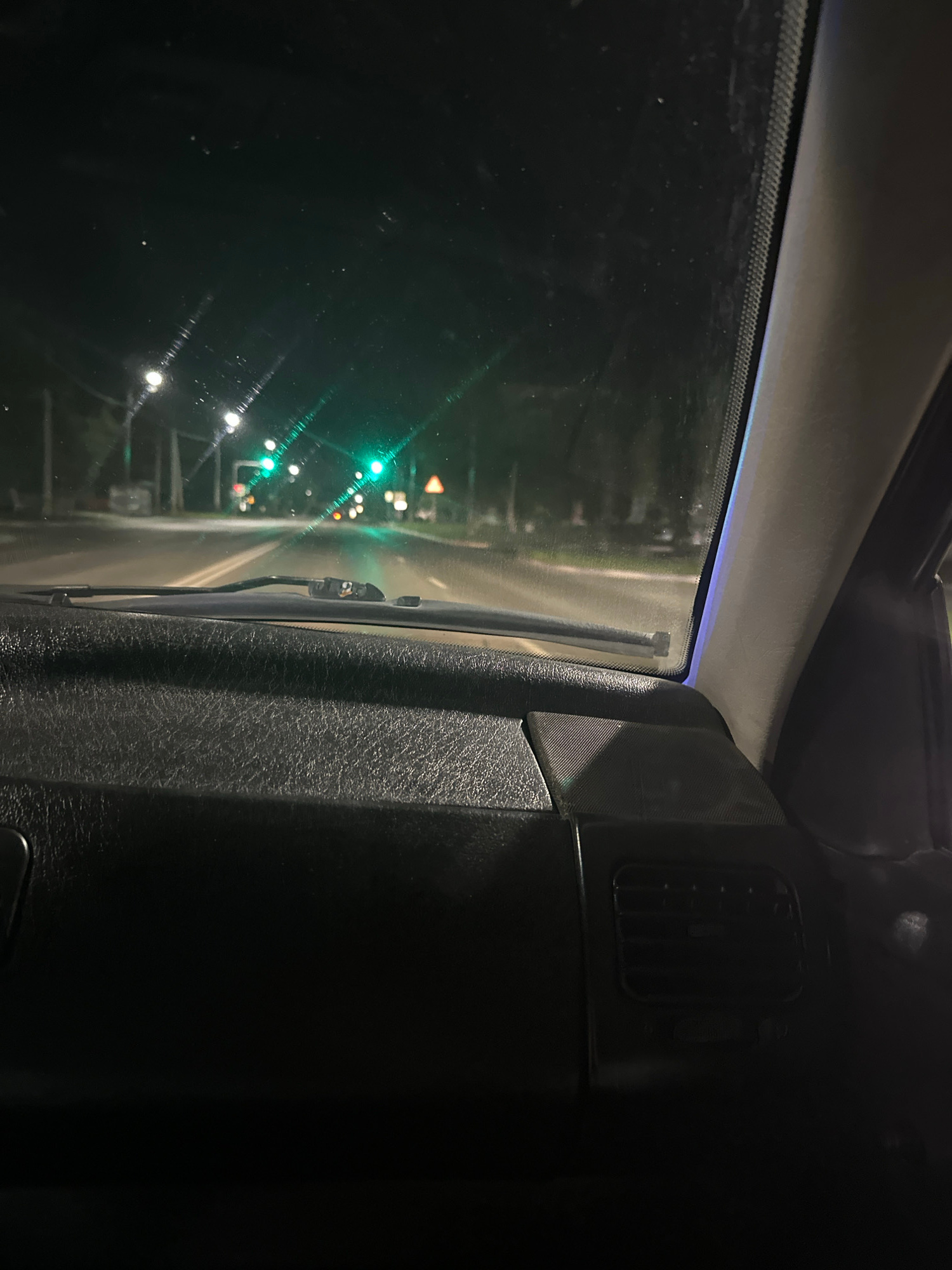 The image shows a view from inside a vehicle, looking through the windshield and out the side window. The time of day is night, as evidenced by the darkness and the visible lights from the road. The scene appears to be a road with traffic lights. The vehicle's interior is partially visible, with dark, textured material on the right side of the frame, possibly the dashboard or door panel. A vent is also visible on the interior panel. The exterior view shows a road with several green traffic lights, suggesting the vehicle is either approaching or at an intersection. Some of the lights are blurred due to motion or the focus of the camera. The weather is not discernible from the image. The location is identified as Kostanay, Kazakhstan. There is no visible text in the image. The overall mood is quiet and possibly reflective, given the nighttime setting and the solitary view from within the car.
