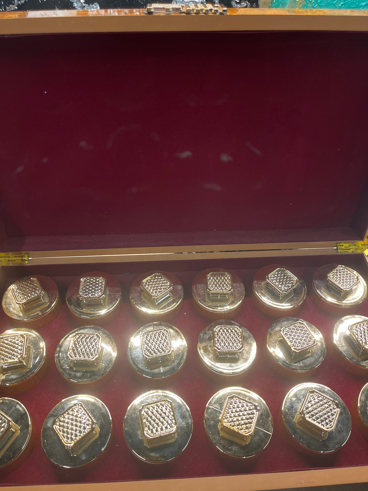 A wooden box with a red interior lining contains multiple gold-colored, round objects. Each object has a raised, square top with a textured, diamond-patterned surface. The box is open, revealing the contents. The image appears to have been taken indoors, possibly in a store or display setting. There are no people or specific landmarks visible that would indicate its location in Krasnodar, Russia. The lighting suggests an artificial light source.