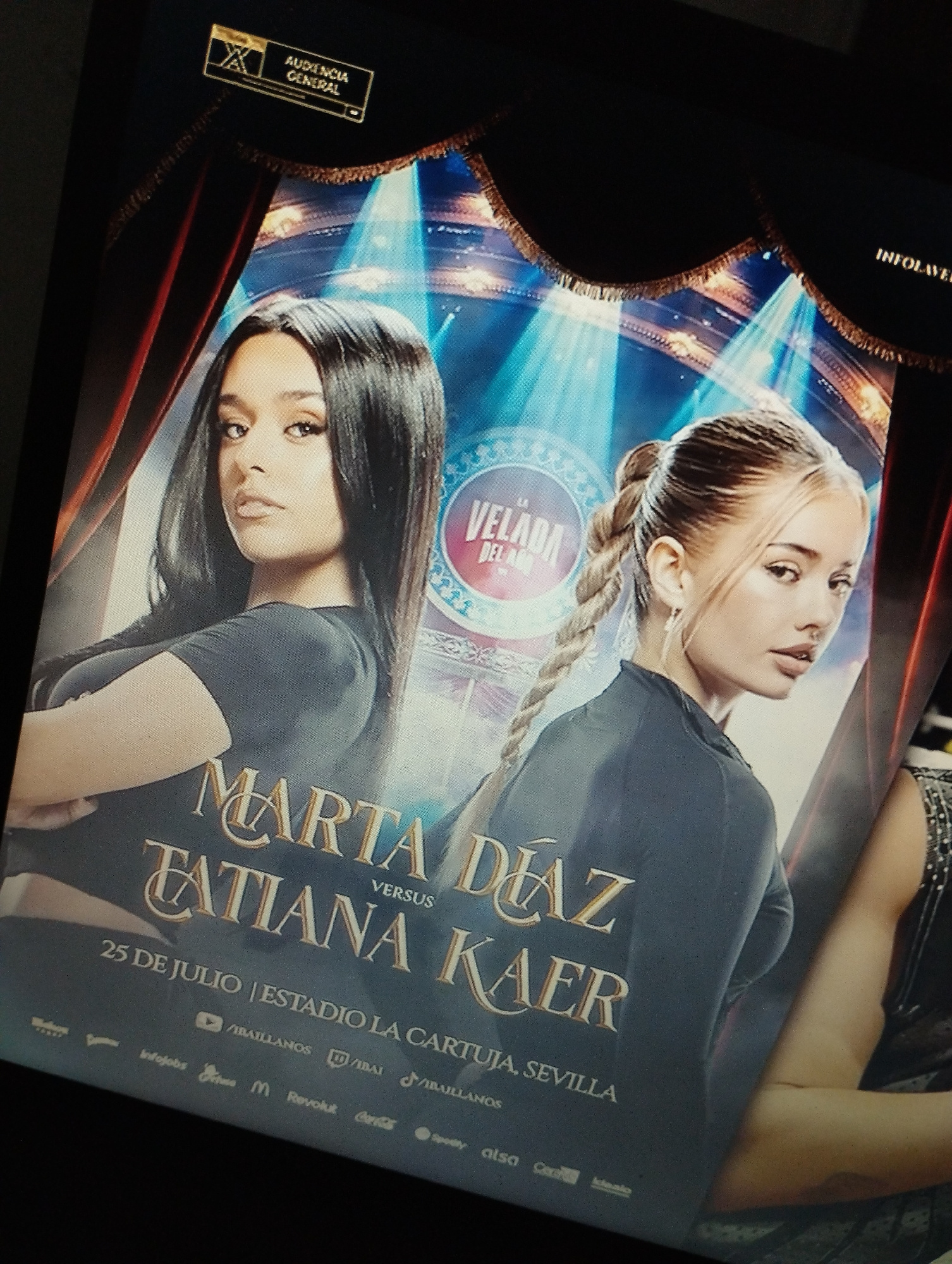 The image is a poster for an event called "LA VELADA DEL AÑO" featuring two young women. The woman on the left has long dark hair and is looking directly at the viewer. The woman on the right has blonde hair styled in a braid and is looking over her shoulder. Both women are wearing dark tops. The poster text reads "MARTA DÍAZ VERSUS TATIANA KAE." Below this, it states "25 DE JULIO | ESTADIO LA CARTUJA, SEVILLA." Several logos of sponsors and social media platforms are visible at the bottom of the poster, including YouTube, Twitch, TikTok, Revolut, and Coca-Cola. The background features theatrical curtains and spotlights. The image is a photograph of a screen displaying the poster. There is no information in the image that places it in Santa Ana, Venezuela.