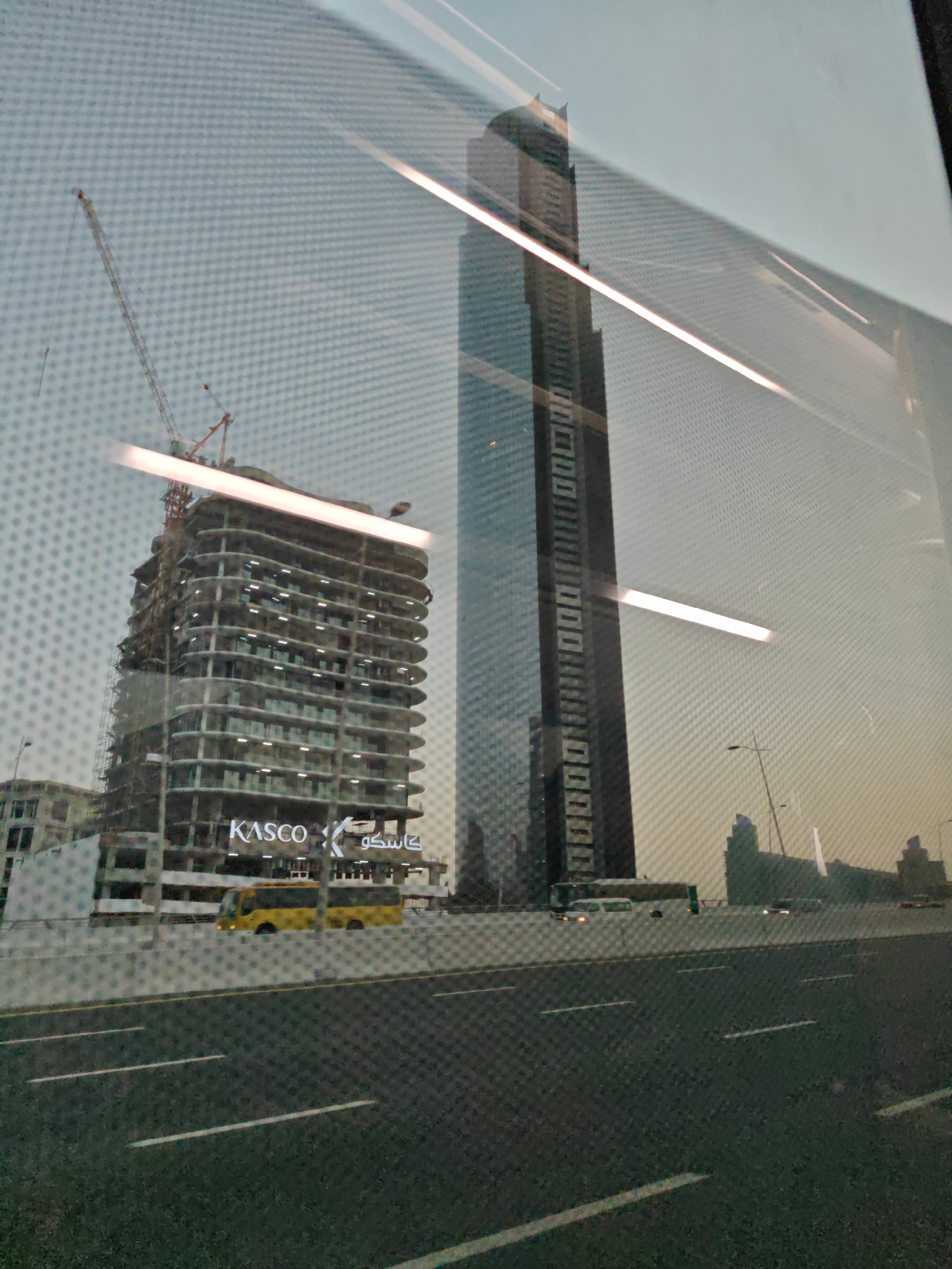 The image shows a reflection on a textured surface, likely glass, capturing an urban scene in Dubai, United Arab Emirates. In the foreground, a highway with white lane markings is visible, with a yellow bus driving on it. Further back, a multi-story building is under construction, with a large crane positioned above it. The building has a distinctive wavy facade and illuminated signage that reads "KASCO" in English and Arabic. To the right of the construction site, a tall, slender skyscraper with dark windows dominates the background. Other buildings and streetlights are scattered throughout the cityscape, suggesting a densely populated urban environment. The sky is overcast, and the lighting indicates it might be late afternoon or early evening.