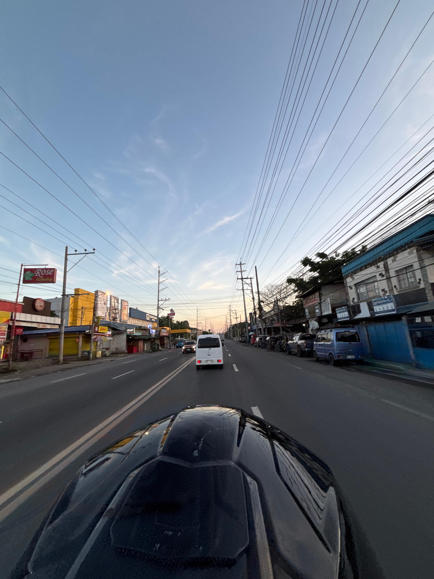 This is a first-person perspective image taken from a motorcycle, looking forward down a street in Davao City, Philippines. The time of day appears to be late afternoon or early evening, as indicated by the soft, warm light and the clear, pale blue sky with wispy clouds. Numerous power lines are strung across the sky, a common sight in Philippine cities.
The road is paved and has clear lane markings. On the left side of the road, there are commercial establishments with colorful signage, including one that reads "Rose." Buildings vary in height and architectural style, with some appearing to be older, single-story structures and others taller, multi-story buildings. There are several parked vehicles on the left side, including motorcycles and a white van.
On the right side of the road, there are more buildings, some with blue accents and metal fences. Several vehicles are also visible on this side, including a blue van and other cars. The overall environment suggests a busy urban street with ongoing commercial activity and traffic. The motorcycle's helmet is prominently visible in the foreground, obscuring the rider's face and lower body.