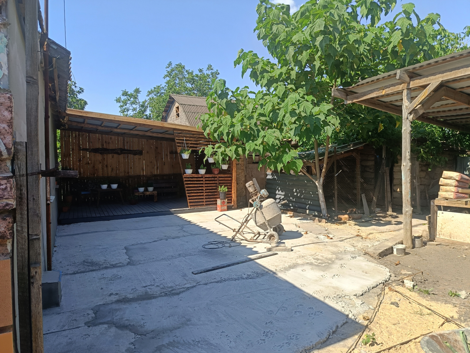 This image depicts an outdoor, rural scene in Peremozhne, Ukraine, likely during the daytime given the bright sunlight and clear blue sky. The setting appears to be a yard or courtyard with elements of construction or renovation in progress.
The main subjects include a concrete mixer, which is centrally placed on the paved ground. A long, narrow wooden plank lies horizontally in the foreground, possibly for mixing or leveling concrete. The ground itself is a mix of concrete and dirt, with some areas appearing freshly poured and others more weathered.
In the background, there is a wooden structure with a sloped, corrugated roof, possibly a shed or a covered patio. This structure has vertical wooden planks for its walls and a wooden deck area with benches. Several white potted plants hang from the structure, adding a touch of greenery. To the right, a large tree with broad green leaves is prominent, casting shadows on the scene. Further to the right, a covered area with a wooden frame and a corrugated roof is visible, with stacks of bagged material (likely cement or similar construction supplies) piled next to it. A wire mesh fence is also visible in the background, suggesting boundaries or enclosures.
The overall impression is one of outdoor work or DIY projects. There are no people visible in the image. The weather appears to be sunny and warm. The presence of the concrete mixer, unfinished concrete areas, and building materials strongly indicates an ongoing construction or repair activity. The scene is utilitarian and functional, with the natural elements of the tree and sky contrasting with the man-made structures and materials. No visible text is present in the image.