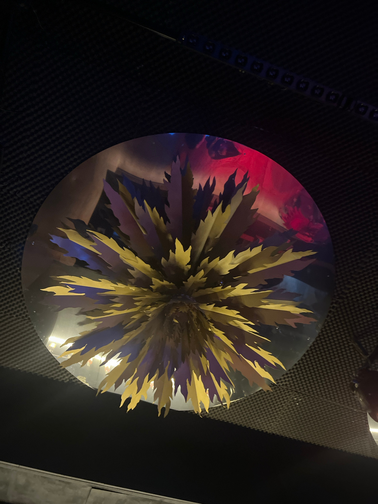 An overhead, close-up, eye-level shot captures a circular, reflective surface, likely a table, with a decorative centerpiece. The centerpiece is a spiky, starburst-shaped object made of layered, pointed yellow and purple paper or cardstock. The reflective surface shows a distorted, abstract reflection of people, with hints of red and blue lighting. The background consists of a dark, textured surface with a grid pattern, possibly acoustic foam or a similar material, and a strip of small, illuminated lights across the top edge. The overall lighting is dim and atmospheric, with colored lights contributing to the mood. The image does not contain enough information to determine a specific city or location.