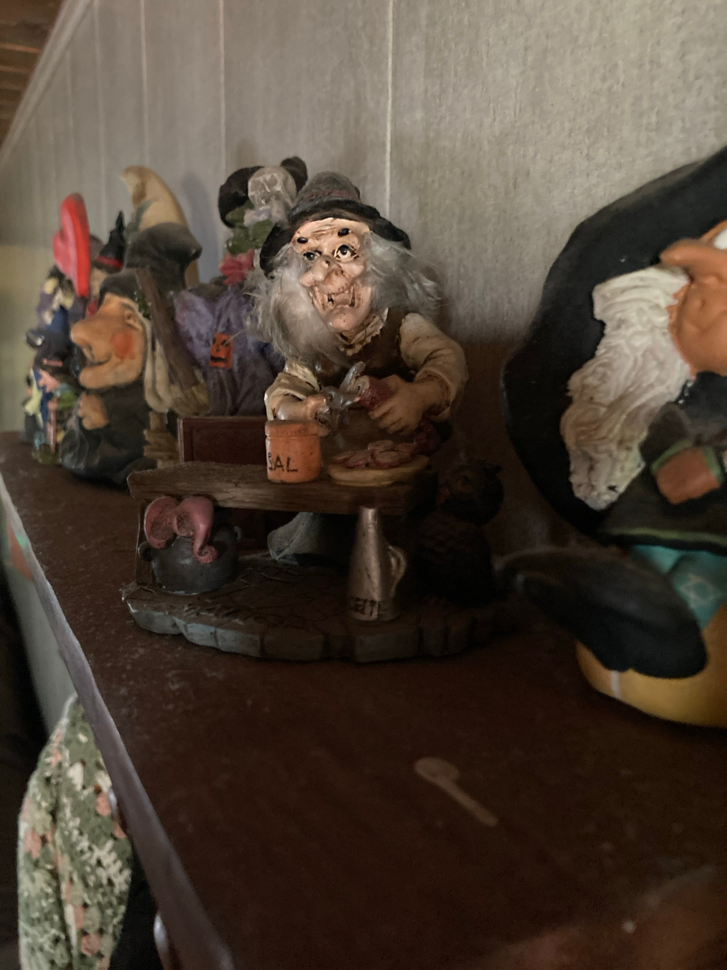 This image depicts a shelf with several decorative figurines, likely for Halloween. The figurines are of witches and gnomes, with one prominent witch in the foreground wearing a hat and an apron, seemingly preparing something on a wooden table. A small orange pot with the word "SAL" (Spanish for salt) is visible on the table. To the left, other figurines are lined up, including one with a red hat and a gnome-like figure. The background shows a textured wall. The image does not contain any elements that would allow for a determination of its location in Sabadell, Spain.