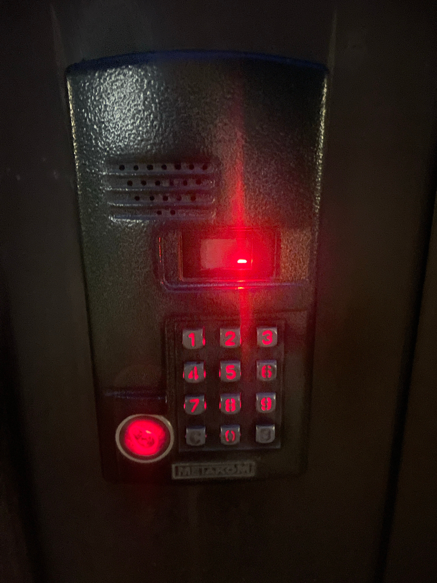 A close-up shot shows a weathered, dark gray intercom panel mounted on a dark background. The panel has a textured surface and features a speaker grille at the top. Below the grille is a rectangular slot with a red light emanating from it. Beneath this are illuminated red numeric buttons arranged in a standard keypad layout, from 1 to 9, with 0 and a star or pound key at the bottom. To the left of the numeric keypad is a circular red button, which is also illuminated. The word "METAKOM" is visible in a silver plate at the bottom of the panel. The overall impression is that of a functional, if somewhat aged, entry system. The location is identified as Markovsky, Russia.