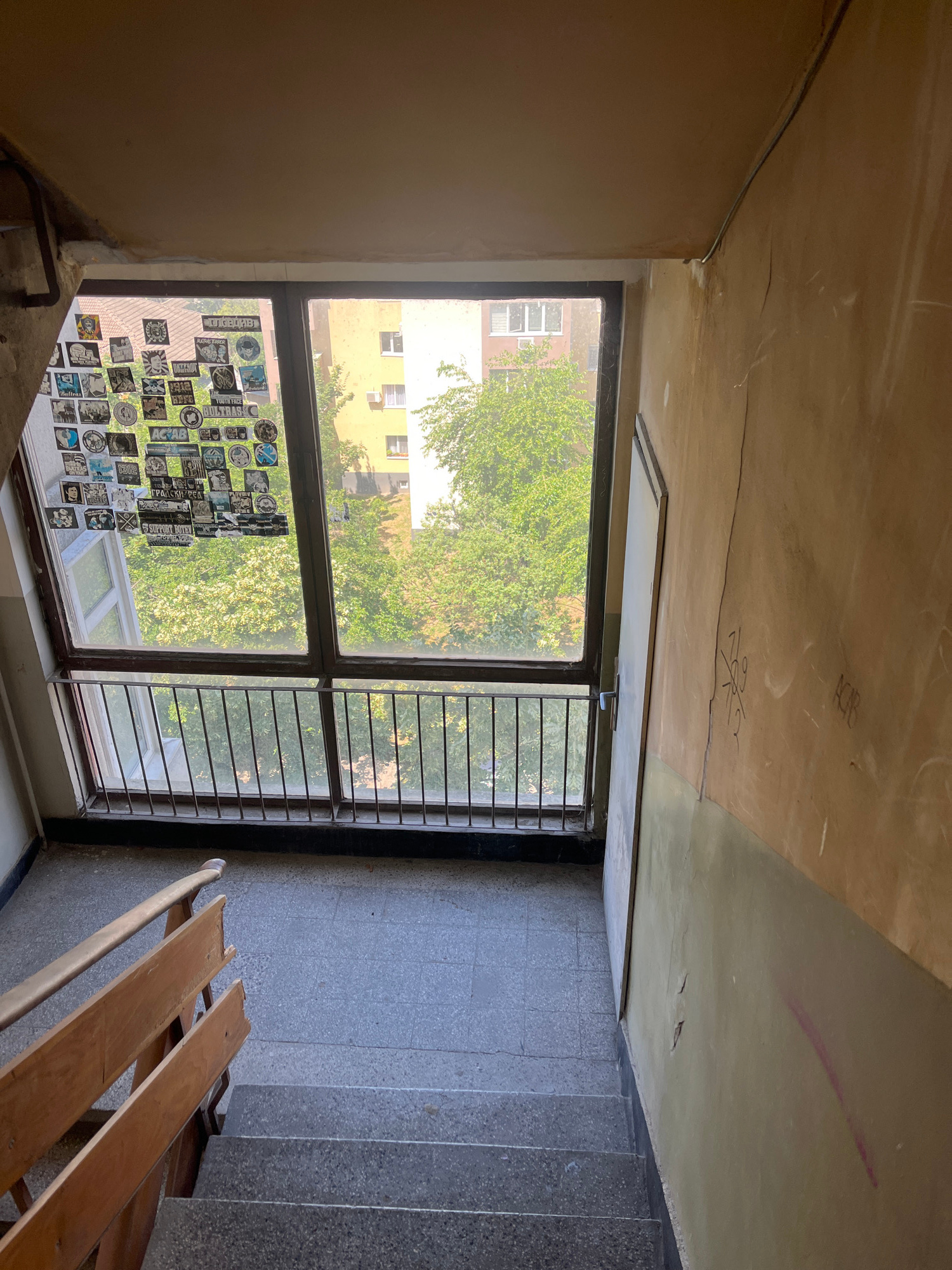 The image shows a view from a staircase landing looking towards a large window and a section of the stairwell. The window is covered with numerous stickers. Outside the window, trees and apartment buildings are visible. The walls of the stairwell are aged and textured, with some visible cracks and graffiti. A wooden handrail and the top of a staircase leading down are also present.