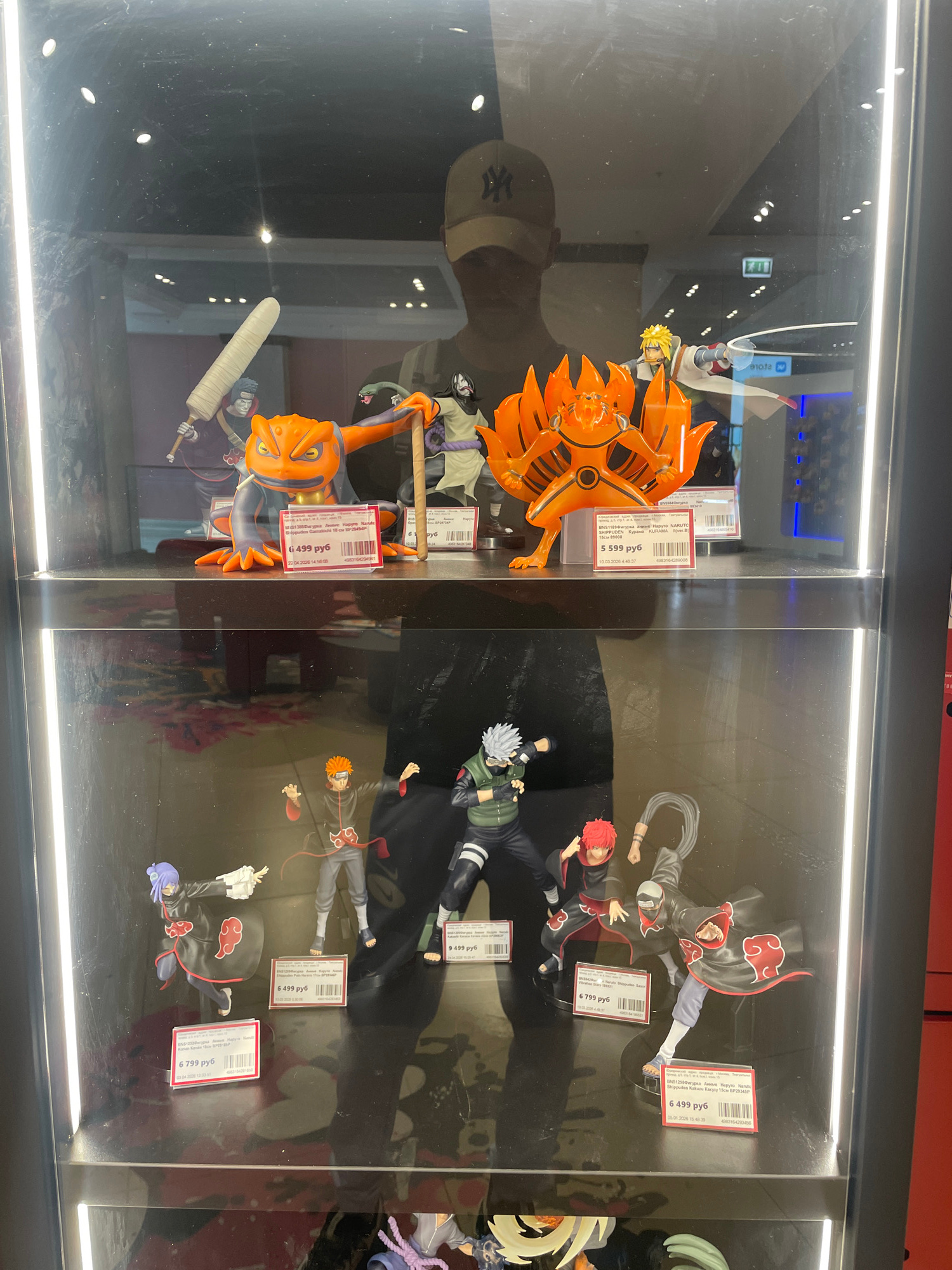 This is a medium shot of an indoor display case, likely in a retail store, showcasing anime figurines. The display is brightly lit from above and the front, causing reflections on the glass surface. The figurines are from the anime series Naruto, with several recognizable characters from the Akatsuki organization, including Kakuzu and Konan, as well as a large orange figure resembling the Nine-Tailed Fox, Kurama.
Several price tags are visible, indicating the cost of the figurines in Russian Rubles (руб). One tag reads "6 799 руб," another "5 599 руб," and another "6 499 руб." The date and time stamps on the tags suggest a recent date. The overall scene is a product display, focusing on collectible items for fans of the Naruto franchise. The figurines are highly detailed, capturing the distinctive designs and poses of the characters. The background is dark, making the brightly colored figurines stand out. The perspective is slightly angled, looking into the display case.