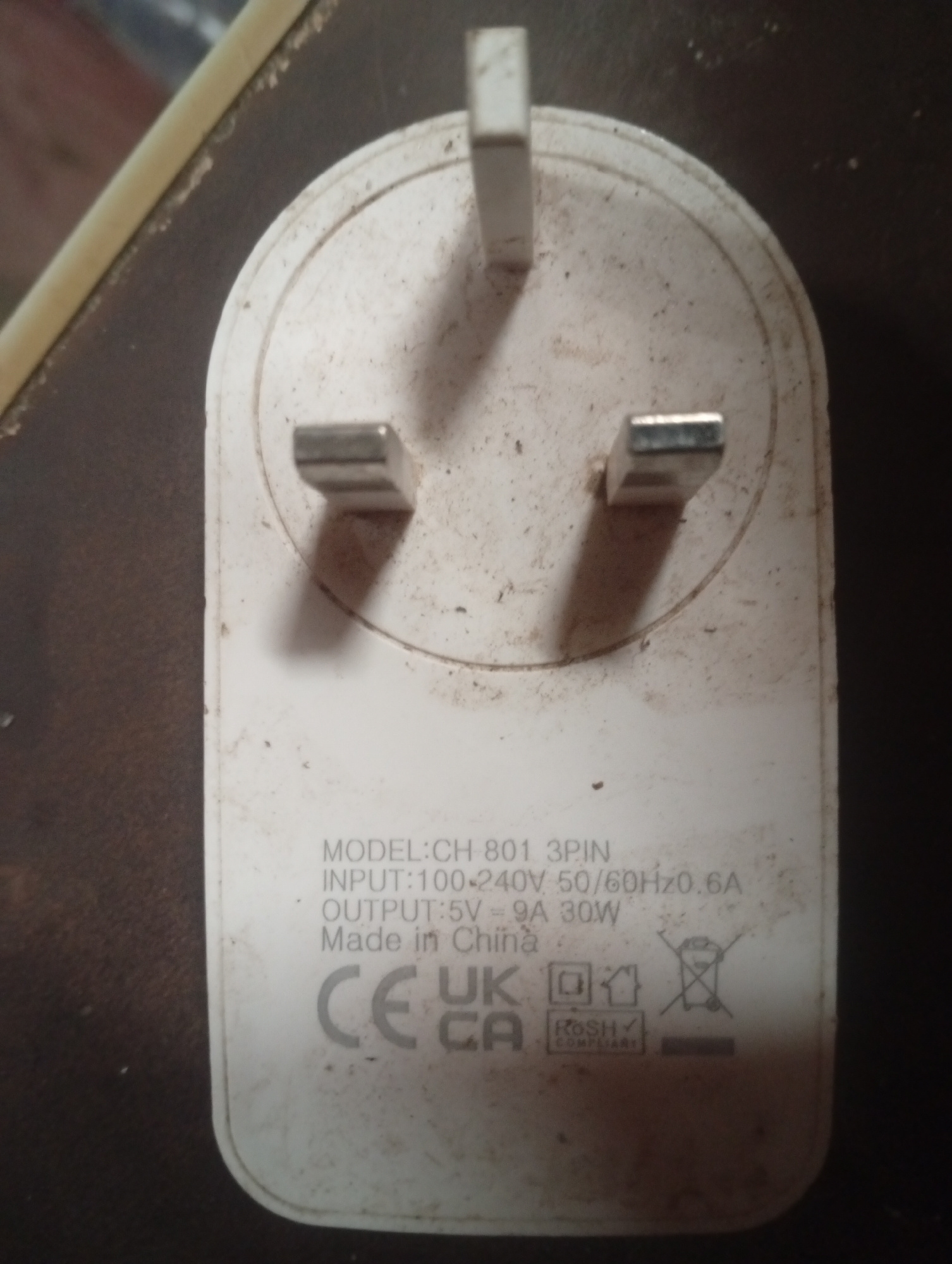 A white, three-pin electrical plug is positioned on a dark, textured surface, possibly a desk. The plug, which appears to be a UK-style plug, is covered in dust and smudges. Markings on the plug indicate it is a "MODEL: CH 801 3PIN," with an "INPUT: 100-240V 50/60Hz 0.6A" and "OUTPUT: 5V = 9A 30W". It also states "Made in China" and displays various certification marks including "CE", "UK CA", a symbol of a power plug inside a square, and a "RoHS COMPLIANT" label. A crossed-out bin symbol is also visible, indicating proper disposal requirements. A thin, light-colored object, possibly a cable, runs along the top left edge of the frame. The lighting in the image is dim, casting shadows from the plug's pins.