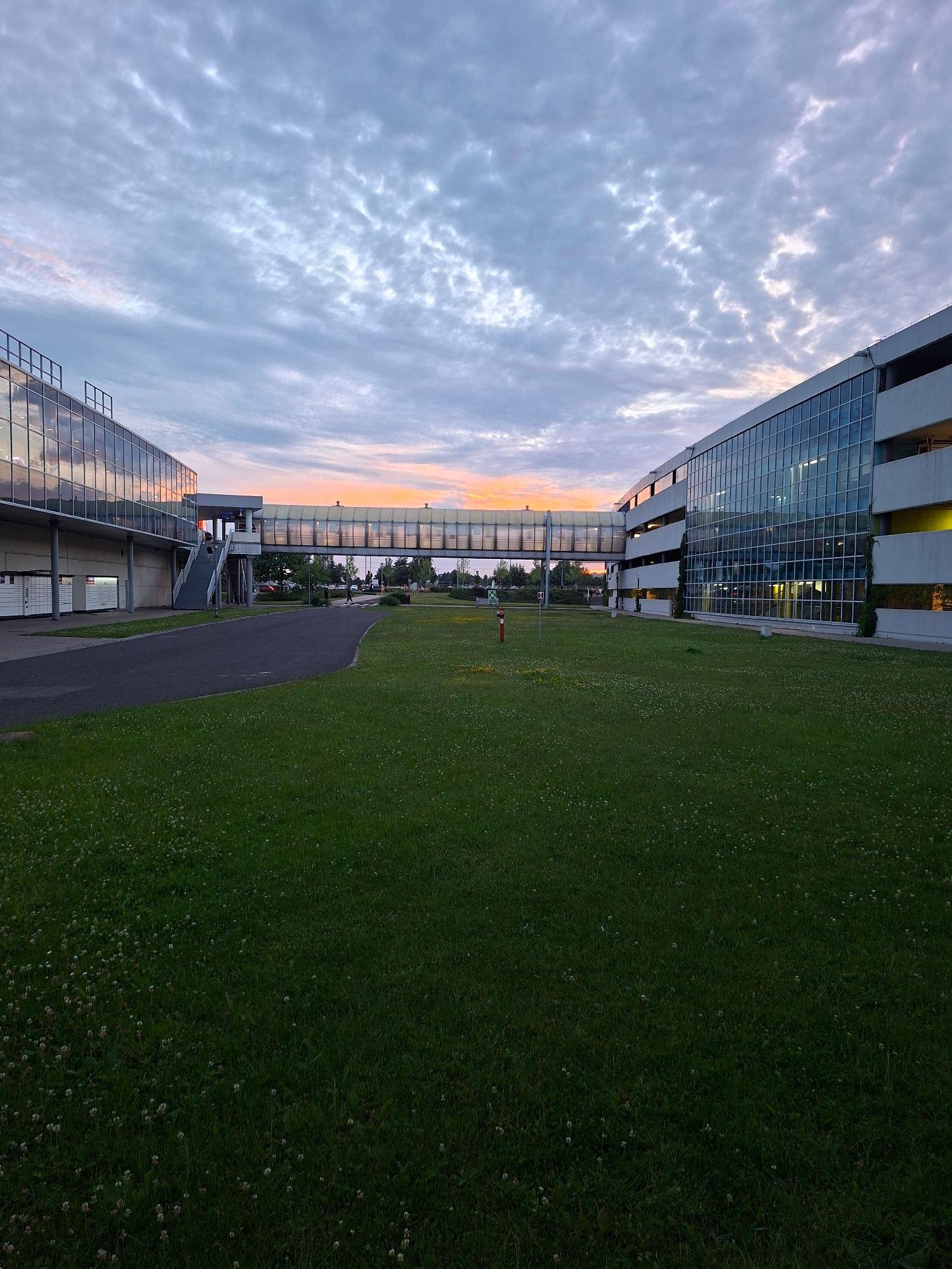 The image shows a modern building complex with a skybridge connecting two sections. The skybridge is a glass-enclosed walkway suspended over a green lawn. The building on the left has a glass facade, reflecting the sky and surrounding structures. The building on the right appears to be a multi-story structure, also with extensive glass windows. The sky is filled with clouds, with hints of sunset colors near the horizon. The foreground consists of a well-maintained green lawn dotted with small white flowers. A paved pathway curves through the lawn. In the distance, trees and other buildings are visible. There are a few individuals walking on the skybridge and on the pathway. A red fire hydrant is visible on the lawn. The scene appears to be an urban or suburban setting, possibly a business park or educational institution. The image was captured in Warsaw, Poland.