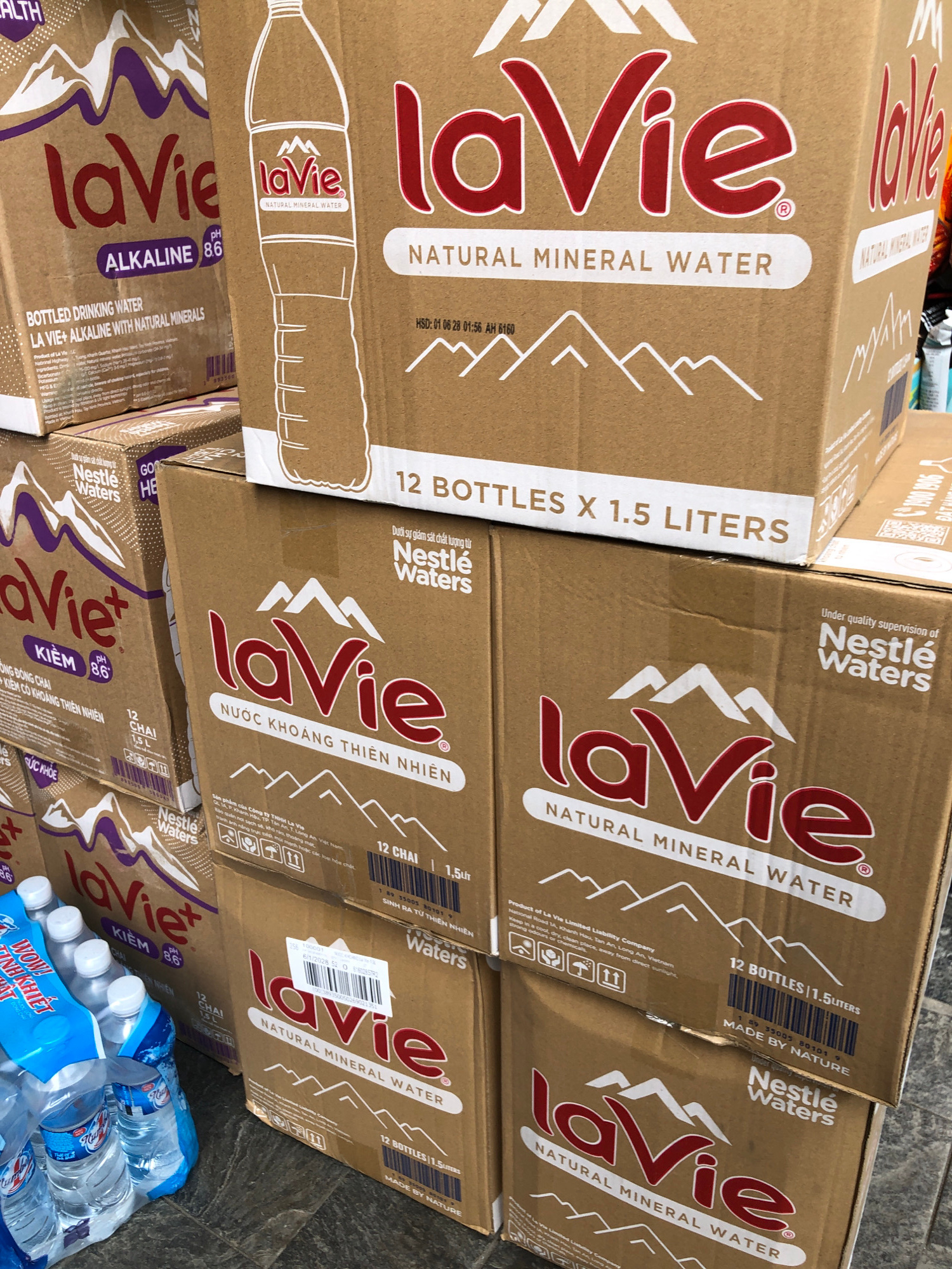 This media file depicts a stack of brown cardboard boxes, likely containing bottled water, arranged on a tiled floor. The boxes are branded with the "La Vie" logo, a well-known bottled water brand in Vietnam, and indicate "Natural Mineral Water." Some boxes specify "12 Bottles x 1.5 Liters" and feature text in both English and Vietnamese, such as "Under quality supervision of Nestlé Waters" and "SINH RA TỪ THIÊN NHIÊN" (Born from Nature). One box highlights "ALKALINE 86" and "LA VIE+ ALKALINE WITH NATURAL MINERALS." The scene appears to be in a storage or retail setting, possibly a warehouse or a shop, given the stacked nature of the products and the tiled flooring. The lighting suggests it could be daytime, possibly indoors given the lack of direct sunlight. There are no people visible in the image. The overall impression is one of product inventory or display.