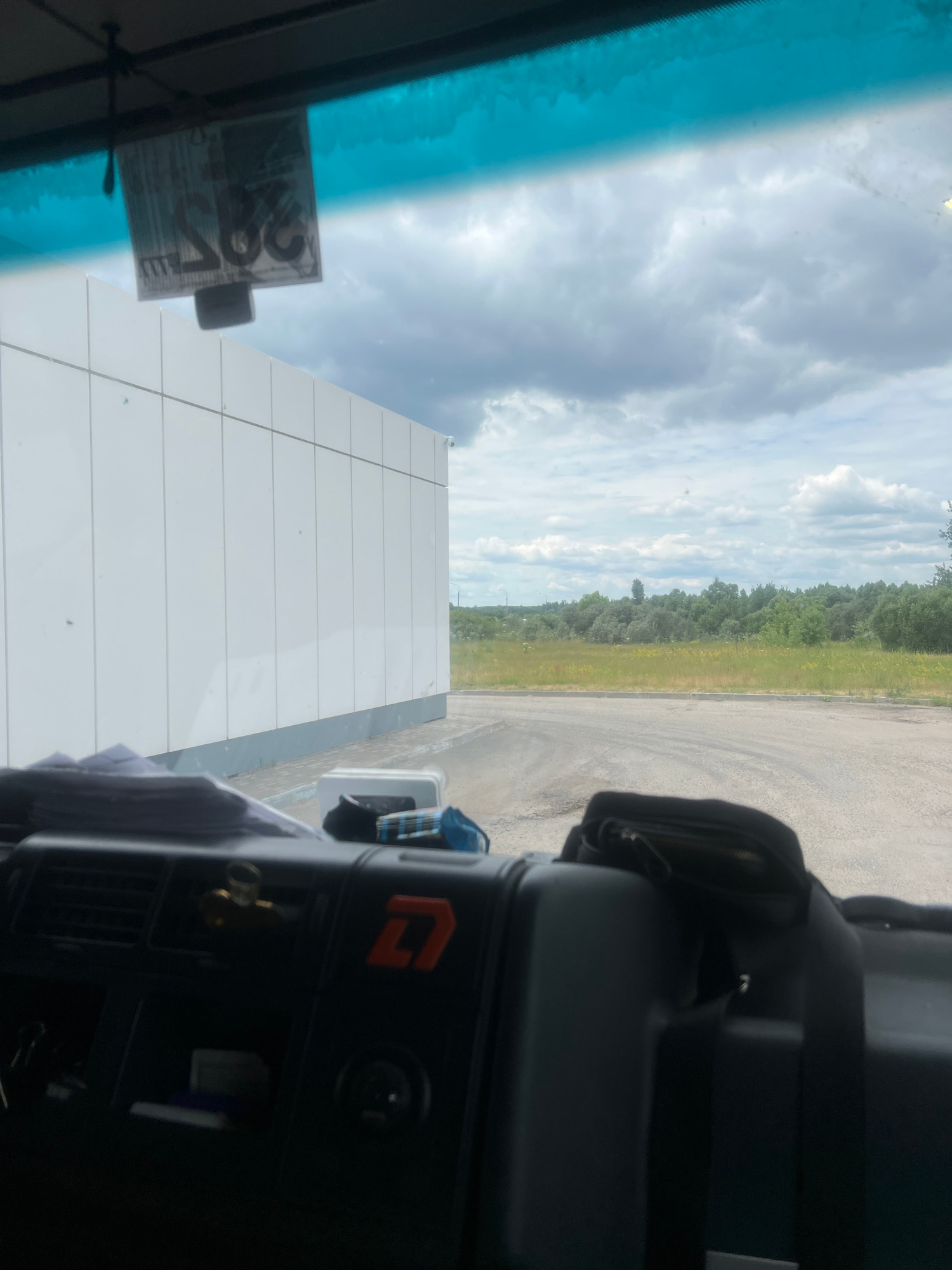 The image is taken from inside a vehicle, looking forward through the windshield. The windshield has a tinted blue band at the top. A tag with Cyrillic lettering and numbers is hanging from the rearview mirror area. To the left, a modern white building with vertical paneling extends across the frame. A small security camera is visible on the side of the building. The ground in front of the building appears to be a paved surface, possibly a road or parking lot, with some water or wetness. Beyond the building and the paved area, there is a wide expanse of green grass and trees under a cloudy sky.
In the foreground, the dashboard of the vehicle is visible. It is black plastic with various vents and controls. A small golden figurine is placed in one of the vents. An orange logo with a stylized "D" is present on the right side of the dashboard. A black bag with a strap is resting on top of some luggage or equipment in the right side of the vehicle's interior. Some small rectangular objects, possibly cards or documents, are visible on the dashboard.
The image does not contain any people. The scene depicts an outdoor environment adjacent to a building, likely in a semi-industrial or commercial area. The location is specified as Chanki, Russia, which would place this scene in a rural or suburban setting in that country.