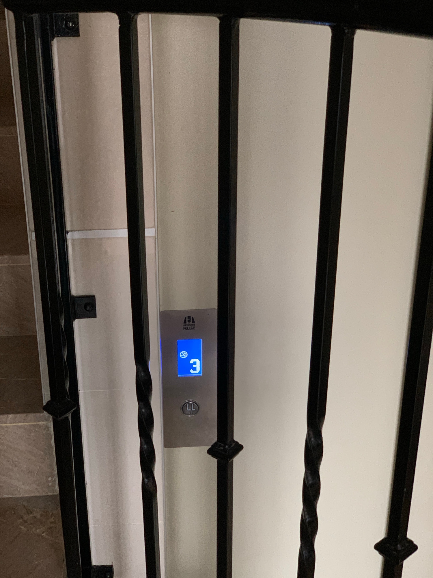 The image shows an elevator control panel with the number "3" illuminated on a blue digital display, indicating the third floor. Above the number, there is a logo and the text "RUIZ." Below the "3" is a button with the inscription "LL." The panel is framed by black metal bars, suggesting it is located in a hallway or stairwell. The wall behind the panel appears to be a light color. The floor tiling visible in the lower left corner is brown and textured. Based on the elevator brand "RUIZ," which is common in Spain, and the overall aesthetic, this scene is likely located in a building in Valencia, Spain.