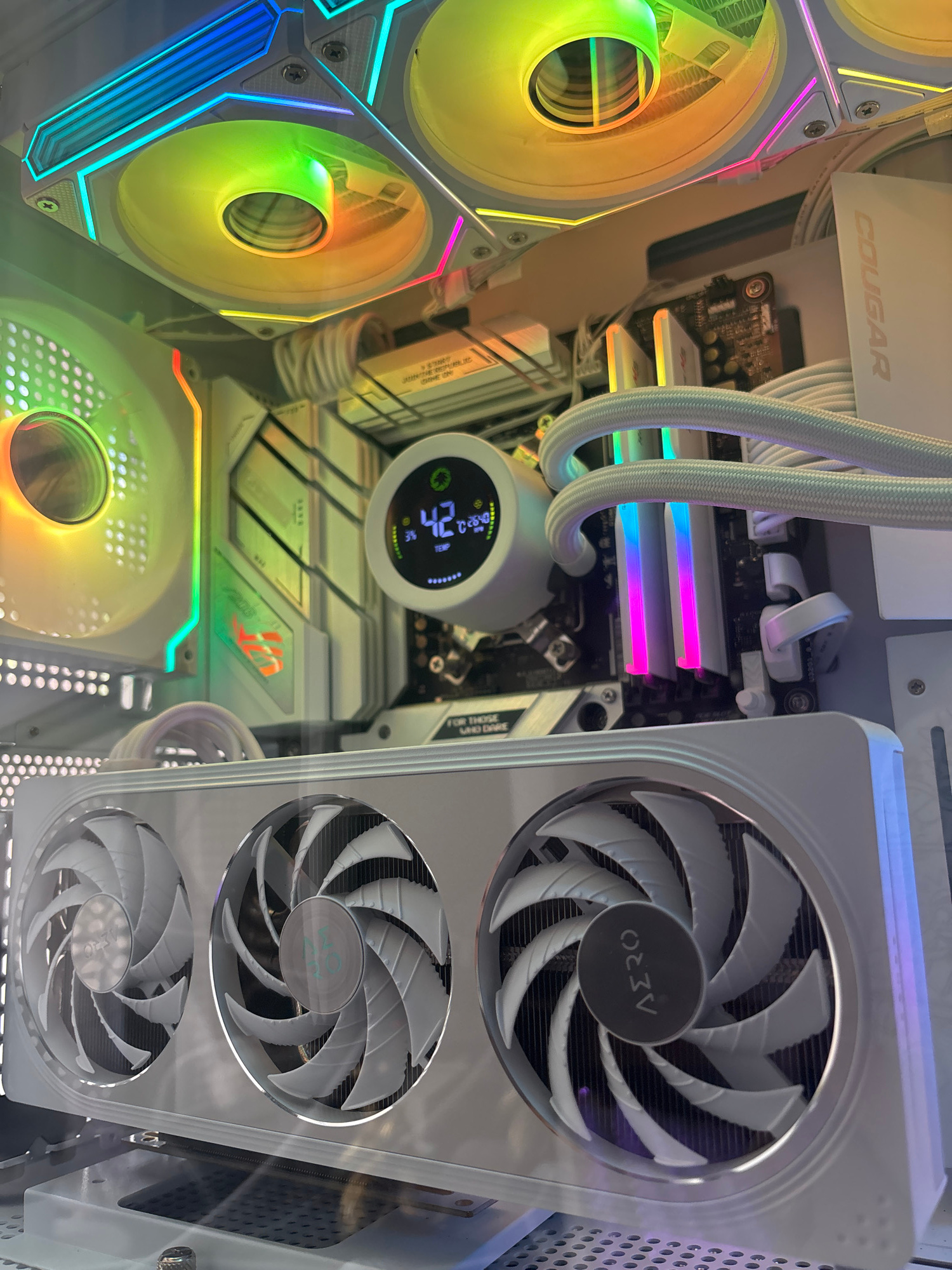 A white computer tower is shown with multiple fans, RGB lighting, and internal components. At the top, two large fans with colorful RGB lighting are visible. Below these, a smaller fan with white blades and RGB lighting is positioned on the left. In the center, a circular LCD screen displays "42" degrees Celsius and other temperature readings. Behind the screen, RAM sticks with pink and blue lighting are installed. The lower portion of the image features a large graphics card with three white fans. The computer tower appears to be placed on a reflective surface, possibly a desk. The overall aesthetic is modern and high-performance, with a focus on white components and customizable RGB lighting.