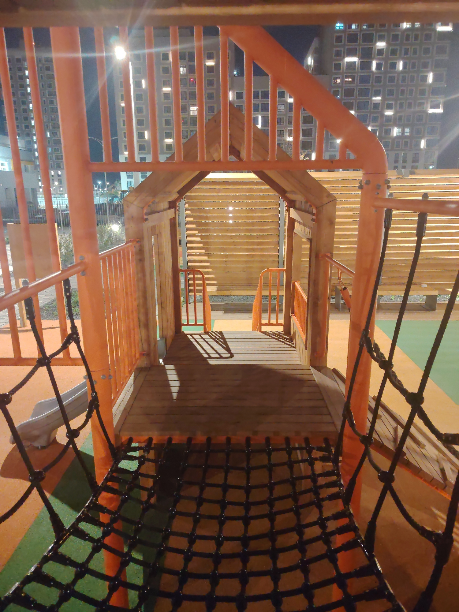 The image depicts a playground structure at night, with artificial lighting illuminating the scene. In the background, tall residential buildings with uniformly lit windows are visible, suggesting an urban environment in Yekaterinburg, Russia. The playground itself is constructed with orange metal bars and wooden elements, including a wooden walkway and a net bridge. There are no people visible in the image. The ground of the playground is a multi-colored surface, with sections of orange and green. The overall impression is of a modern, well-maintained urban playground.