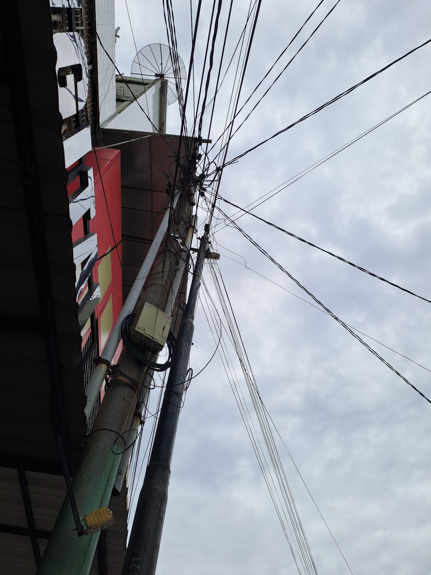 The image captures a low-angle view of a utility pole laden with a dense network of electrical and communication wires against a cloudy sky. To the left, a portion of a building is visible, featuring a red facade with a sign containing white lettering, partially obscured by shadows and the tangle of wires. A satellite dish is mounted on the roof of the building. The utility pole itself is concrete and appears weathered, with a beige electrical box attached to it, from which numerous cables emanate. The overall scene suggests an urban environment with exposed and somewhat chaotic infrastructure. The location is identified as Bandarbireuen, Indonesia, implying a likely residential or commercial area in that city. No individuals are visible in the frame.