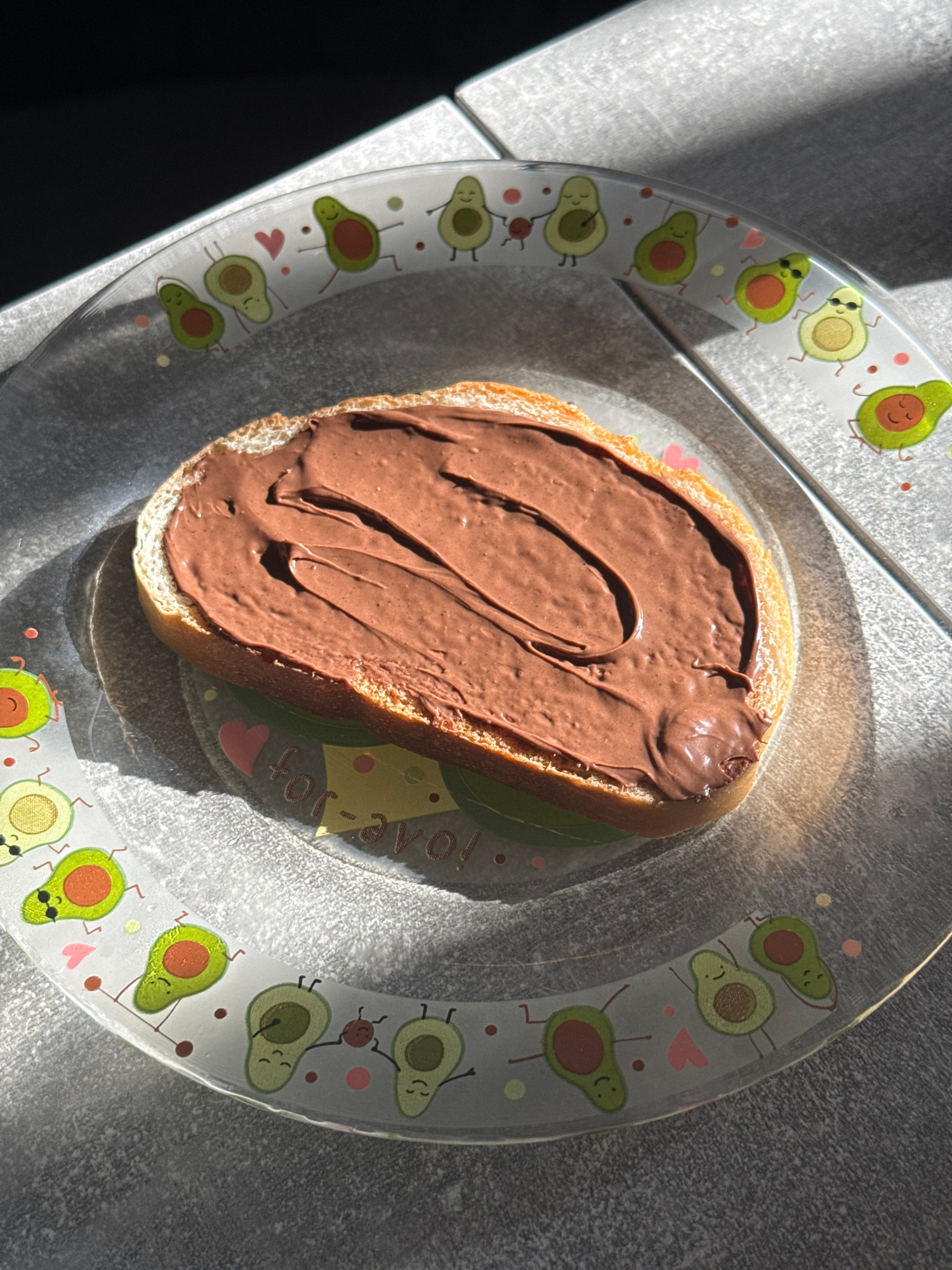 A slice of bread, generously spread with chocolate-hazelnut spread, rests on a clear glass plate adorned with cartoon avocados. The plate is positioned on a textured grey surface, possibly a countertop or table. Sunlight casts distinct shadows across the scene, indicating it's outdoors or near a window. The image is framed to focus on the toast and the plate's decorative edge. No specific location in Lyubertsy, Russia can be determined from the visual information provided.