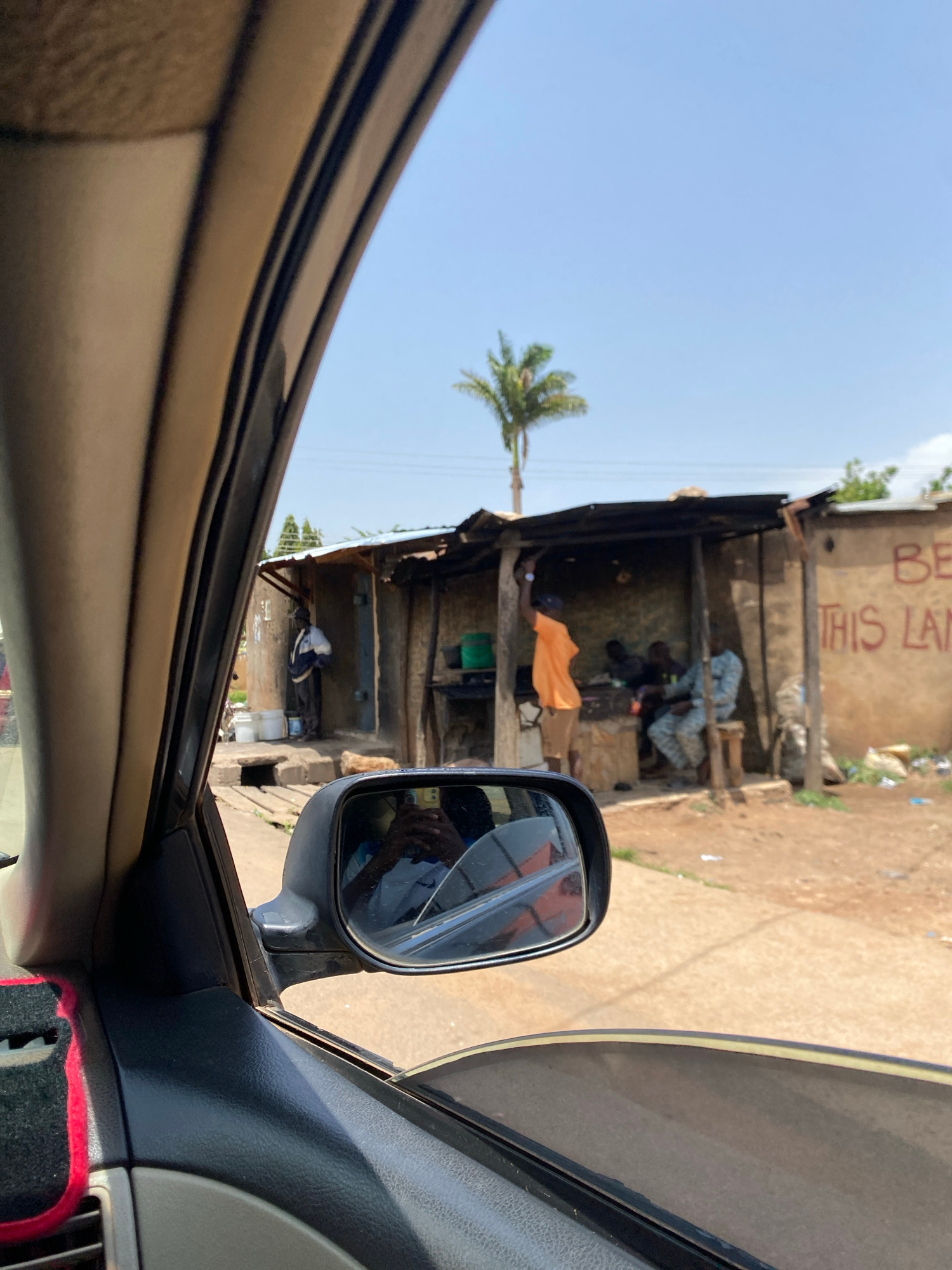The image captures a view from inside a car, looking out through the passenger side window and rearview mirror. In the foreground, the car's interior is visible, including a portion of the dashboard and the side mirror. The side mirror reflects the interior of the car and the photographer. Outside the car, a dusty road stretches ahead, leading to a cluster of small, weathered structures that appear to be shops or stalls. These structures have corrugated metal roofs and are built from various materials, including what looks like mud or concrete. In front of one of the structures, several individuals are present. One person, wearing an orange shirt, is standing near the entrance. Inside, at least two other individuals are seated, and another person in a patterned uniform stands further back. A palm tree is visible in the background, under a clear blue sky. The wall of one of the structures has red lettering that appears to say "BE THIS LAN". The scene suggests a roadside setting in a potentially rural or semi-urban area.