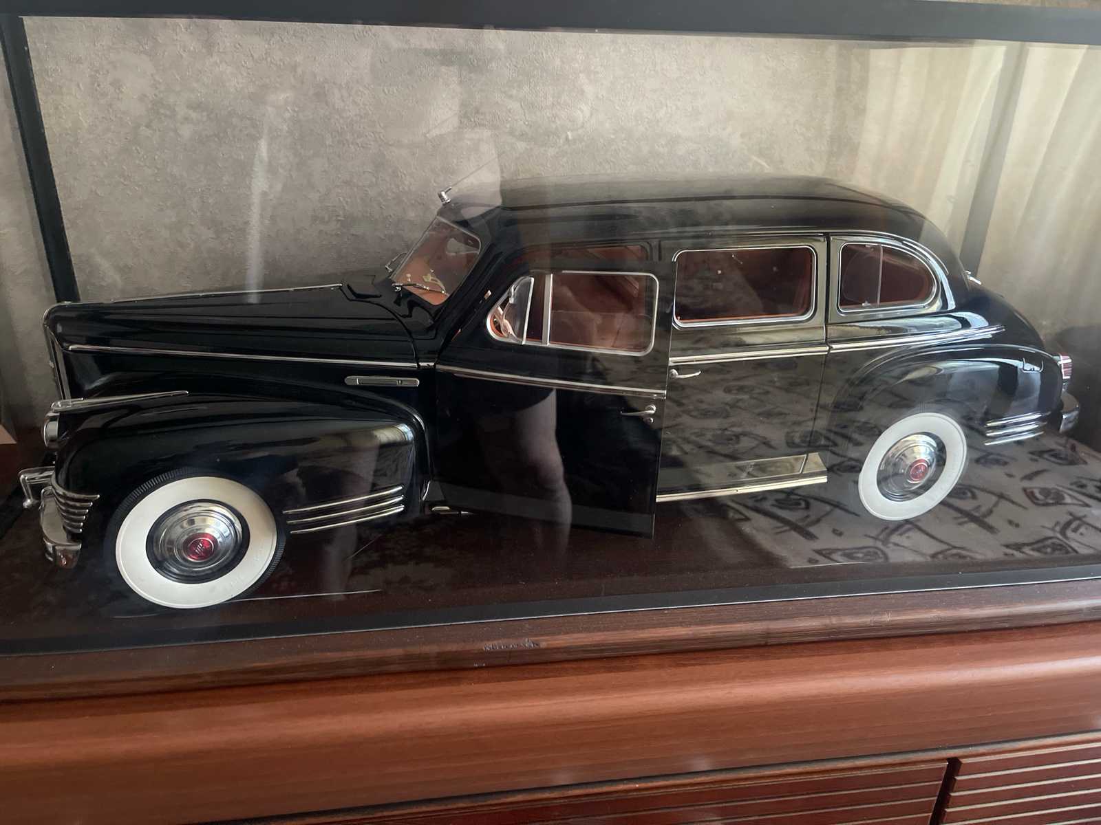 A black 1940s-style sedan model is displayed inside a glass case. The car has whitewall tires with chrome hubcaps, a chrome grille, and a polished black exterior. The driver's side door is open, revealing a tan interior. The car is positioned on a reflective surface, possibly a mirror, which shows a distorted pattern. The background is a textured, light-colored wall, and to the right, sheer white curtains are visible. The car model is placed on a dark wooden surface, likely a piece of furniture, which is in the foreground. The lighting is soft, suggesting an indoor environment, and there are no visible people or explicit indications of time of day or weather. The overall scene appears to be a display of a collectible item. No visible text is present on the car or in the immediate surroundings. The context of Minsk, Belarus is not directly observable from the image itself.
