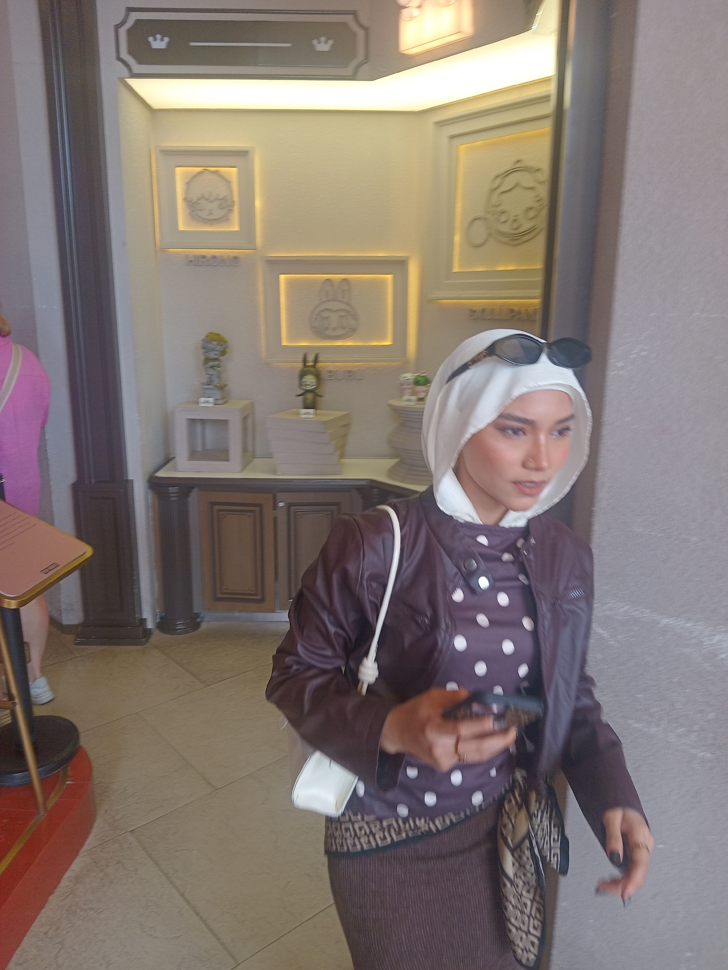 The image captures a woman wearing a white hijab, sunglasses perched on top, a maroon polka-dot shirt, and a matching maroon jacket. She is holding a smartphone in her right hand and has a white purse slung over her left shoulder. The background features decorative frames on a wall, some with figures inside, and display shelves with figurines and books. The setting appears to be an indoor retail or exhibition space, possibly in Da Nang, Vietnam, given the context. The flooring is tiled.