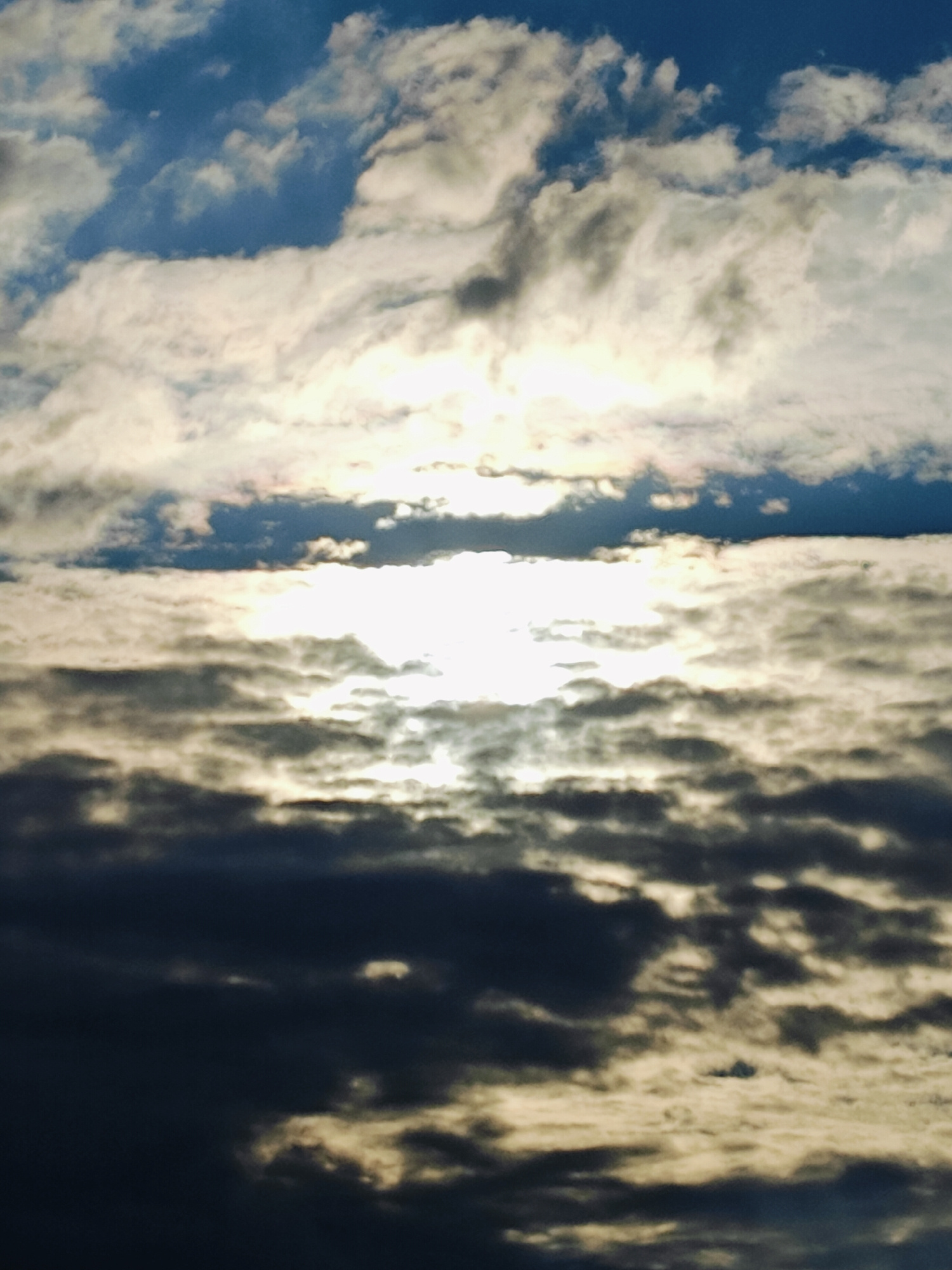 The image displays a dramatic sky filled with a dynamic array of clouds. The sun is partially obscured by these clouds, creating a luminous effect where light filters through, casting bright highlights and deep shadows. The clouds range in color from dark, almost black masses in the foreground to lighter, wispy formations tinged with yellow and white where the sun's rays are most direct. In the upper portion of the image, patches of clear blue sky are visible, suggesting a transition in weather. The overall scene evokes a sense of grandeur and possibly a change in atmospheric conditions, perhaps hinting at an approaching storm or the aftermath of one. The lighting suggests it is either early morning or late afternoon. There is no visible text.