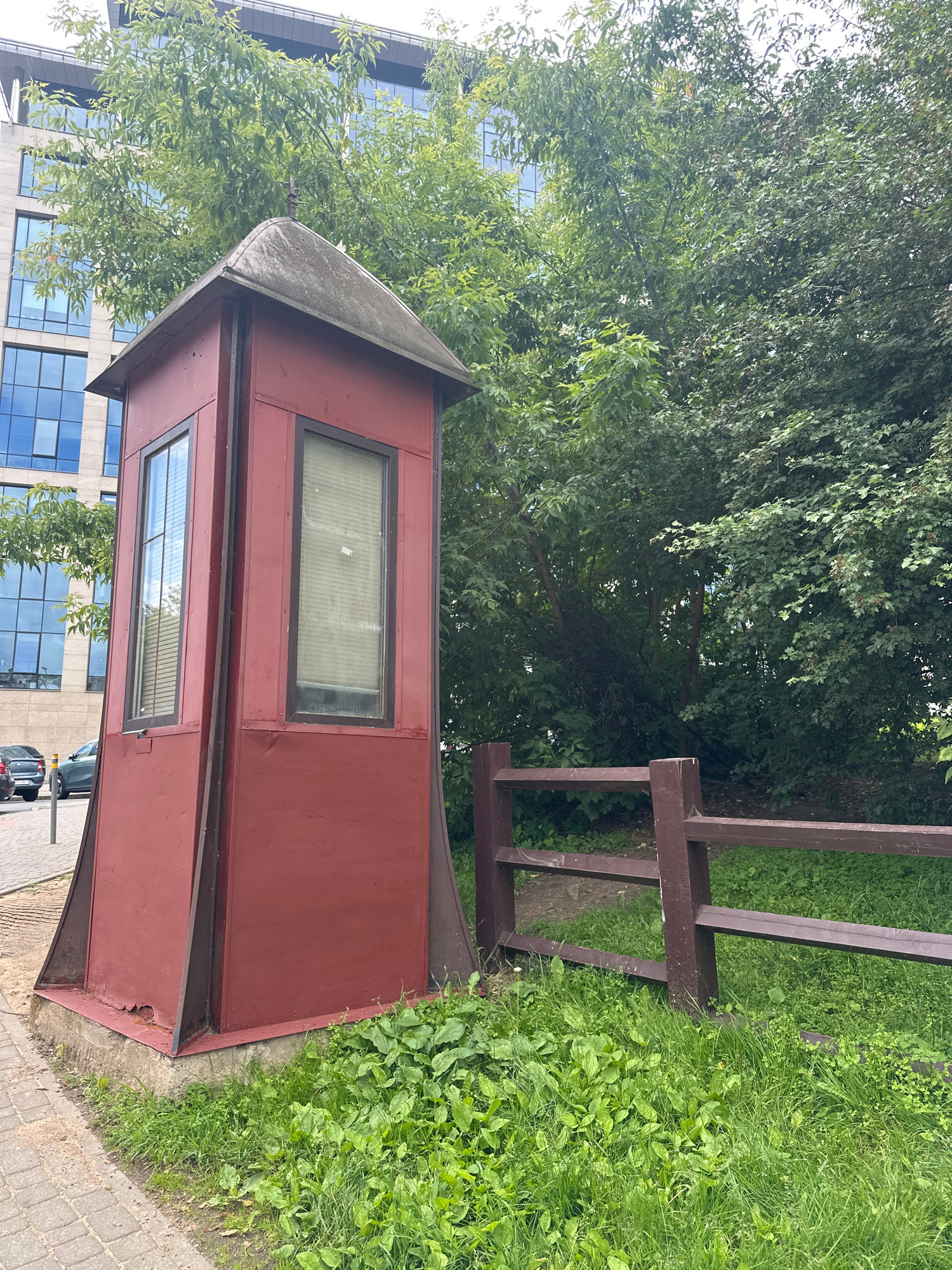 An exterior, eye-level, medium shot captures a small, red, hexagonal booth situated on a paved walkway, adjacent to a grassy area and a wooden fence. The booth features two windows, one with blinds, and a pointed roof. Behind the booth, a modern glass-fronted building with multiple stories and trees are visible. Several cars are parked on the left side of the image, near the walkway. The overall scene depicts an urban setting in Minsk, Belarus.
